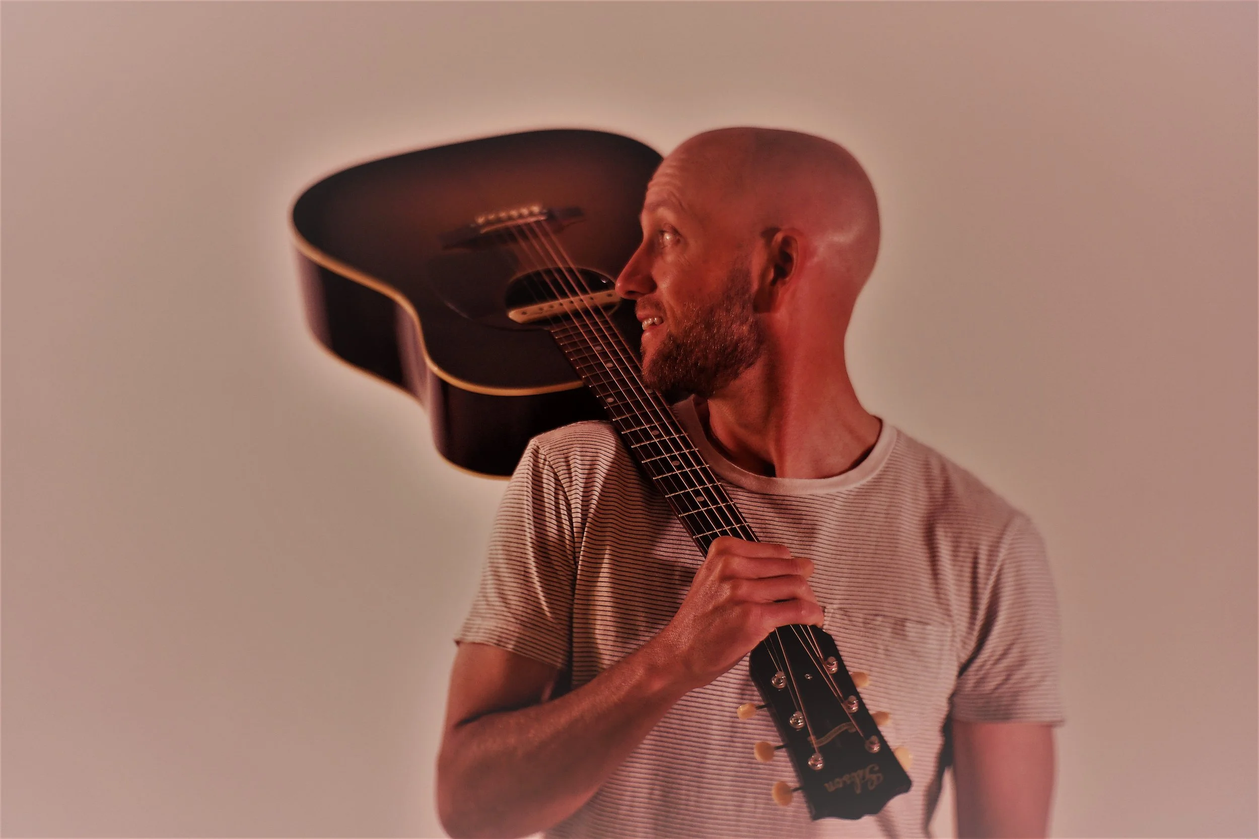 Man with a shaved head and beard, wearing a beige striped t-shirt, holds a black acoustic guitar over his shoulder, looking to the right.