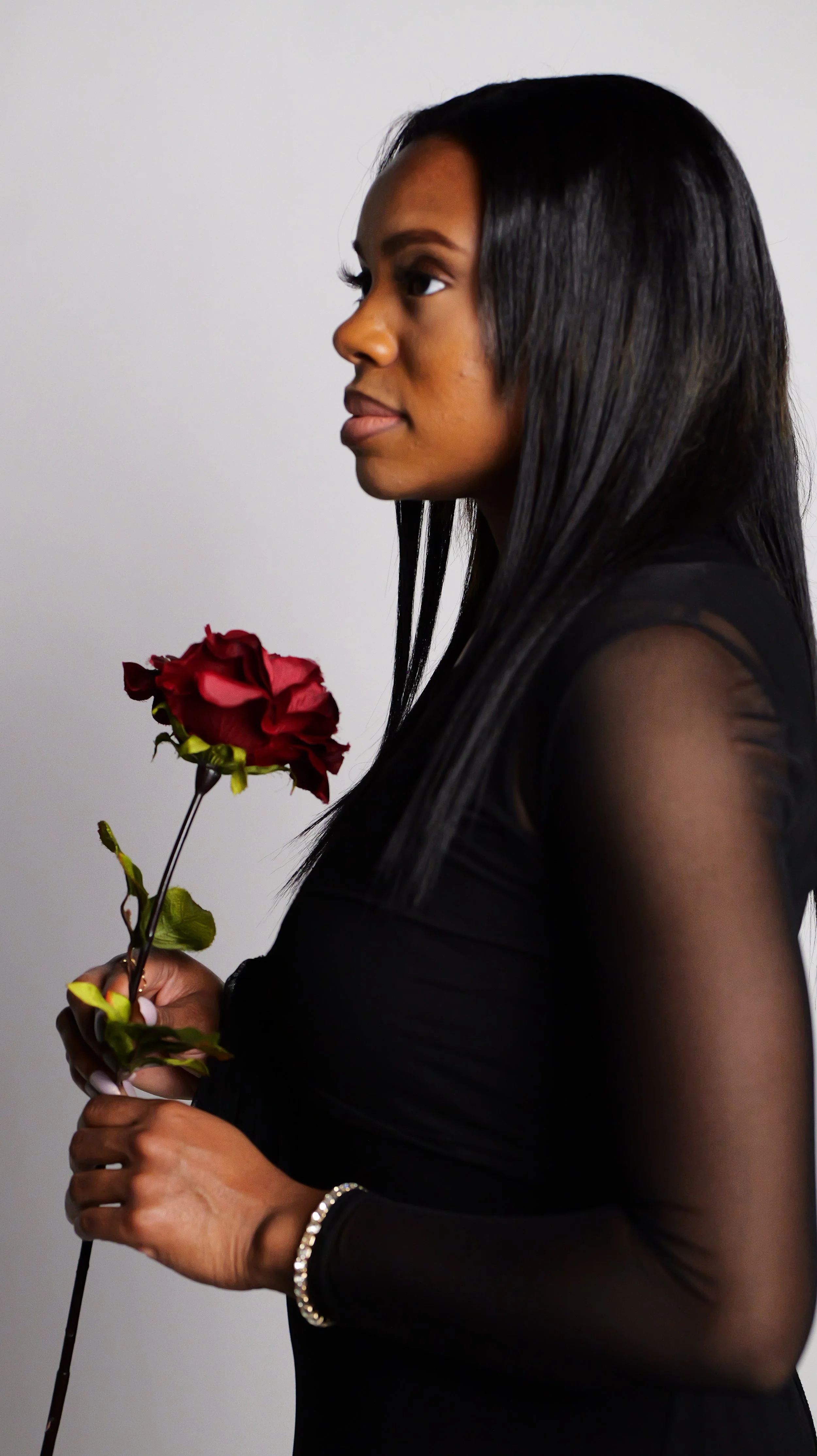 A woman with long black hair and dark skin, wearing a black sheer long-sleeve top, holding a single red rose, standing against a plain background.