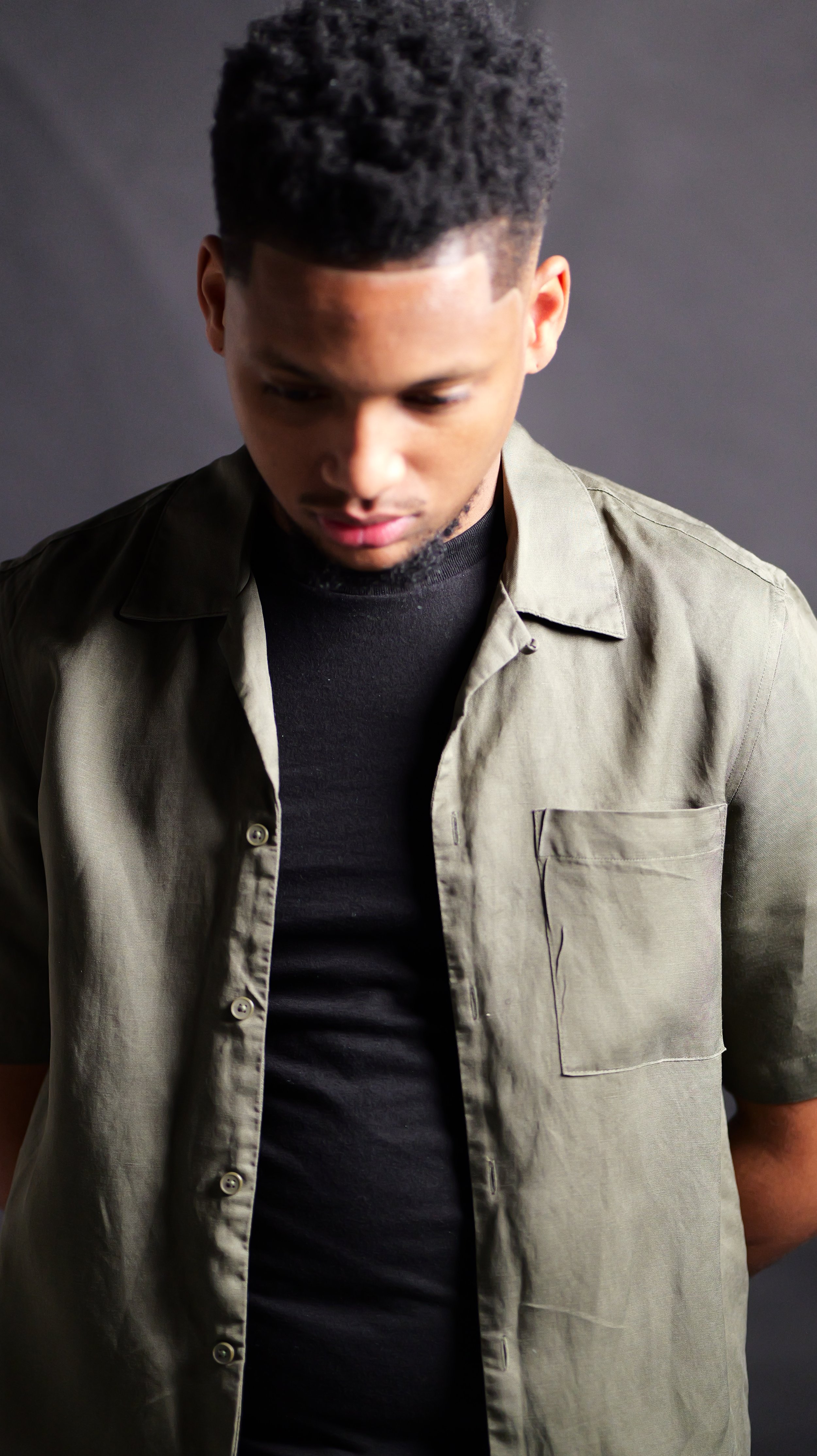 A young man with short curly hair looking down, wearing a black shirt and an open olive-green button-up shirt, against a dark gray background.