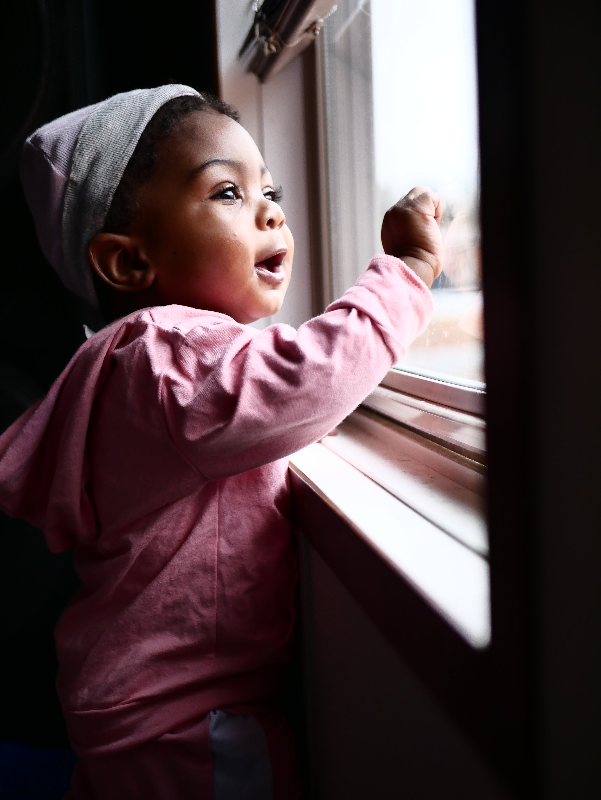 A young girl in a pink hoodie and gray beanie looking out and reaching at a window with sunlight shining through.
