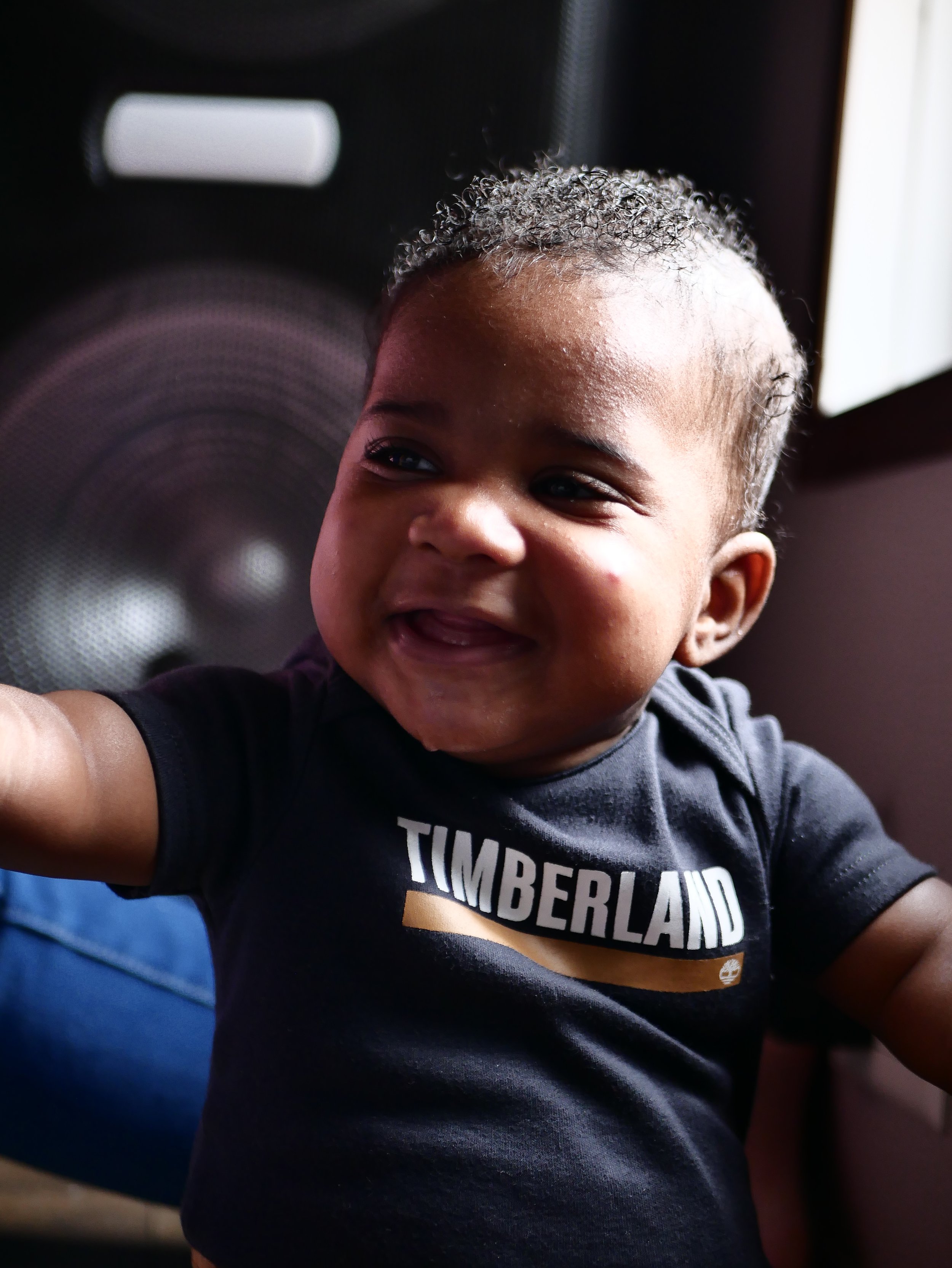 Close-up of a smiling toddler with curly hair, wearing a black T-shirt with the word 'TIMBERLAND' on it, indoors near a speaker and window.