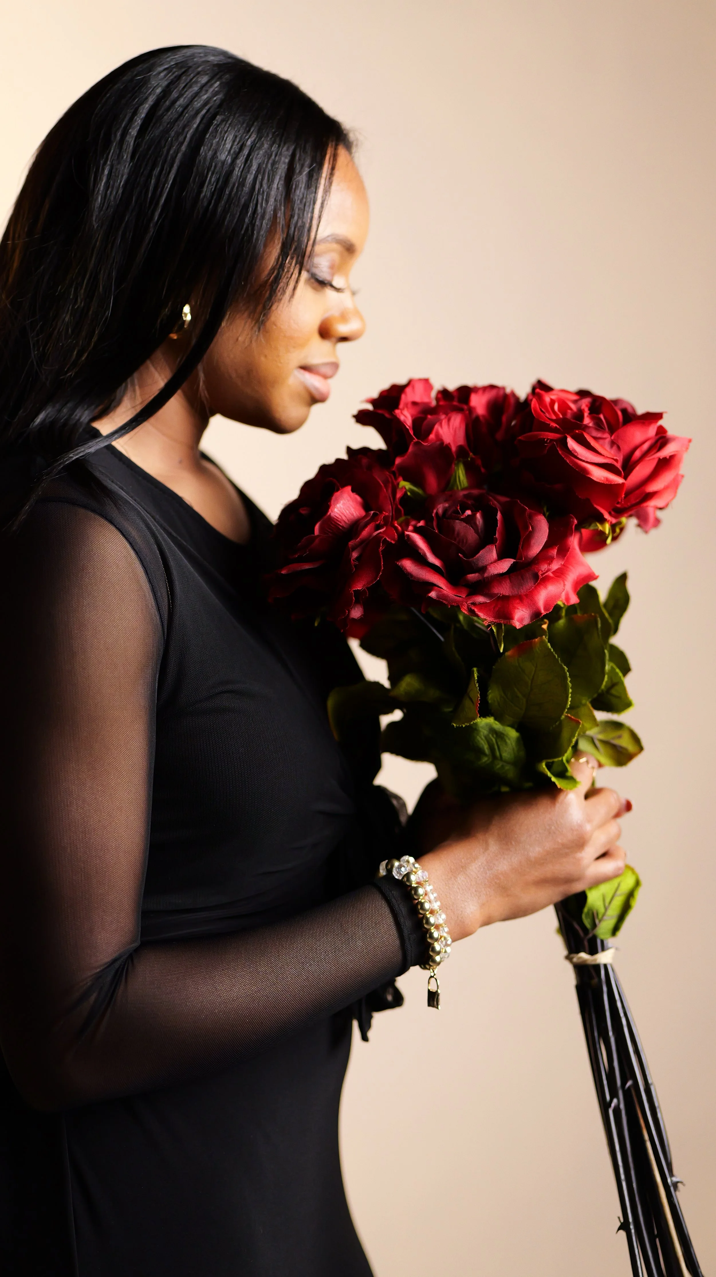 A woman with dark hair in a black dress holding a bouquet of red roses, standing in front of a plain beige background.