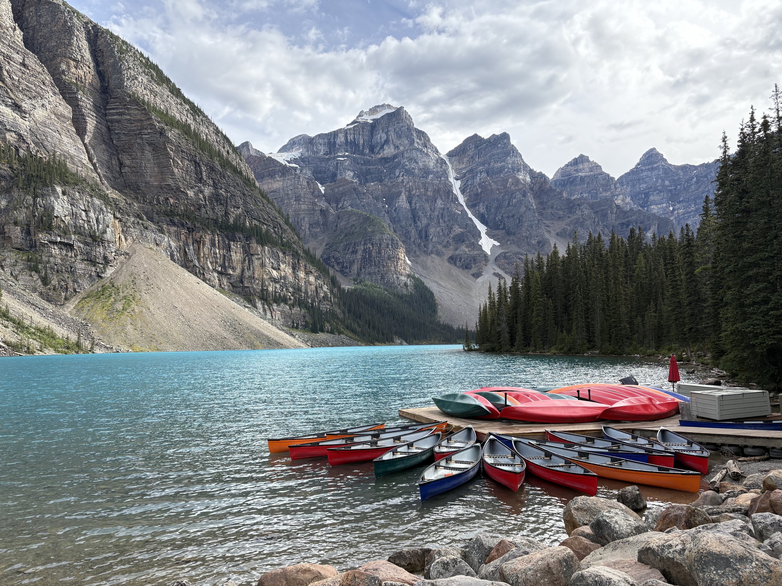 Colorful canoes and kayaks on a dock by a turquoise lake surrounded by mountains and pine trees.