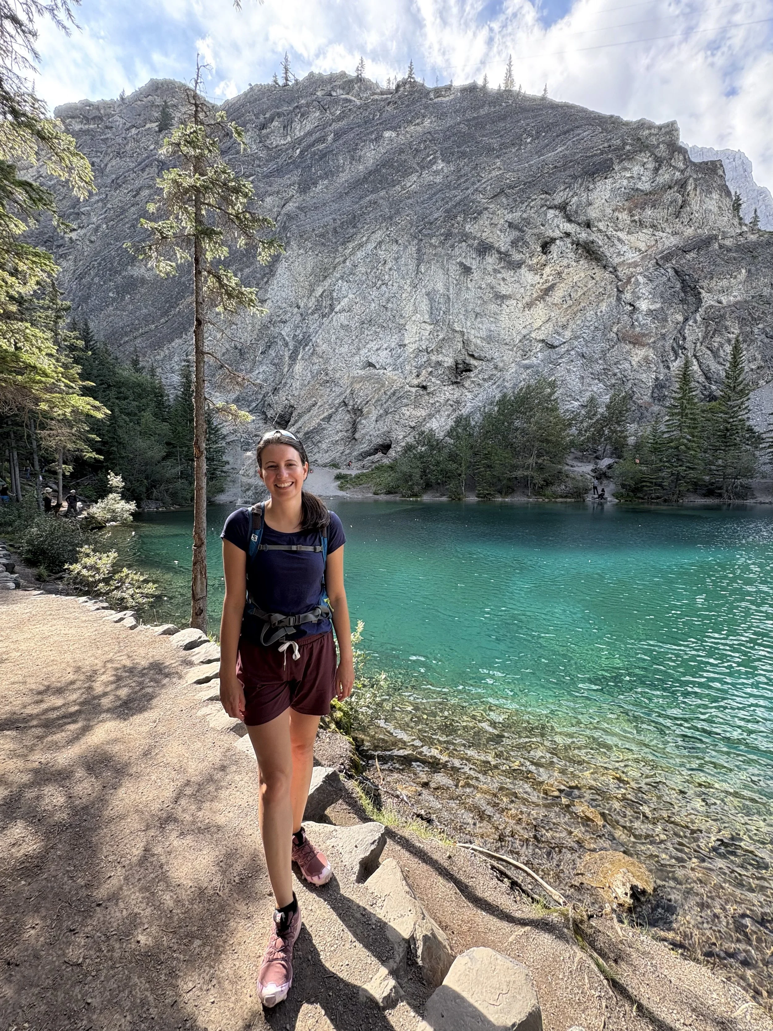 A woman poses by a lake surrounded by trees and rocky mountains, wearing a blue shirt, maroon shorts, and hiking boots.