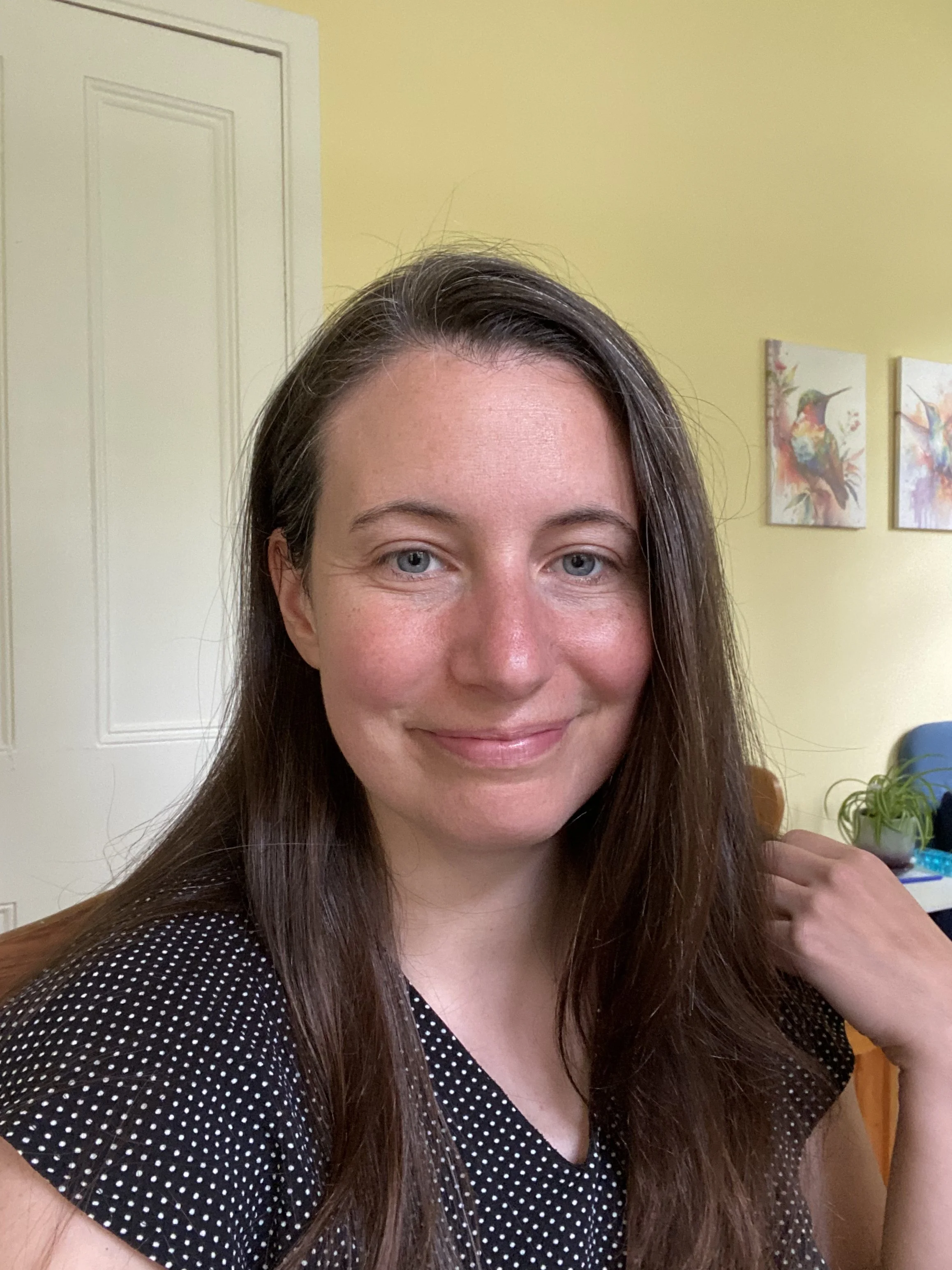 A woman with long brown hair, blue eyes, and a slight smile, wearing a black shirt with white polka dots, sitting in a room with yellow walls, two colorful bird paintings on the wall, a white door, a blue chair, a small table with a green plant, and some miscellaneous items.