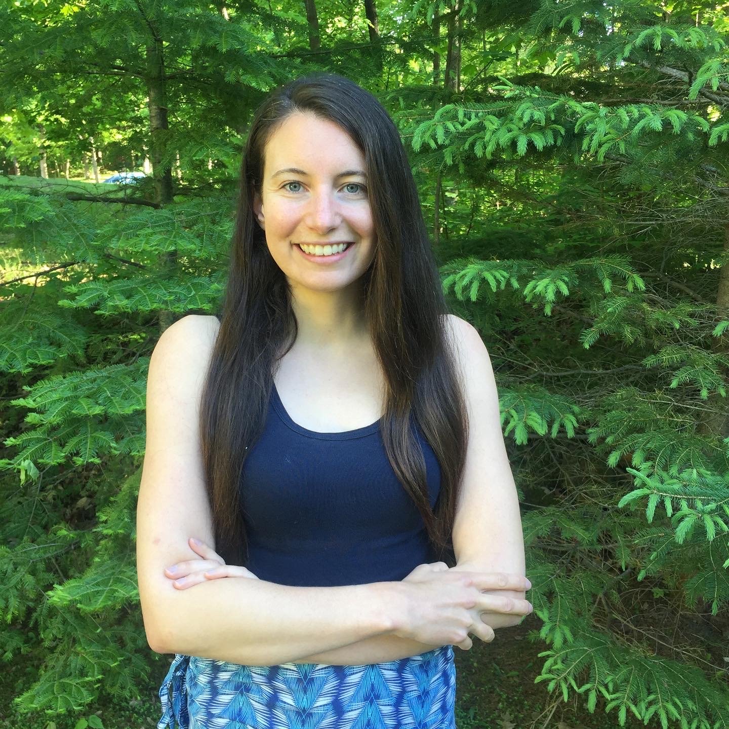 A young woman with long dark hair, wearing a black tank top and blue patterned shorts, standing outdoors in front of lush green trees, smiling with her arms crossed.