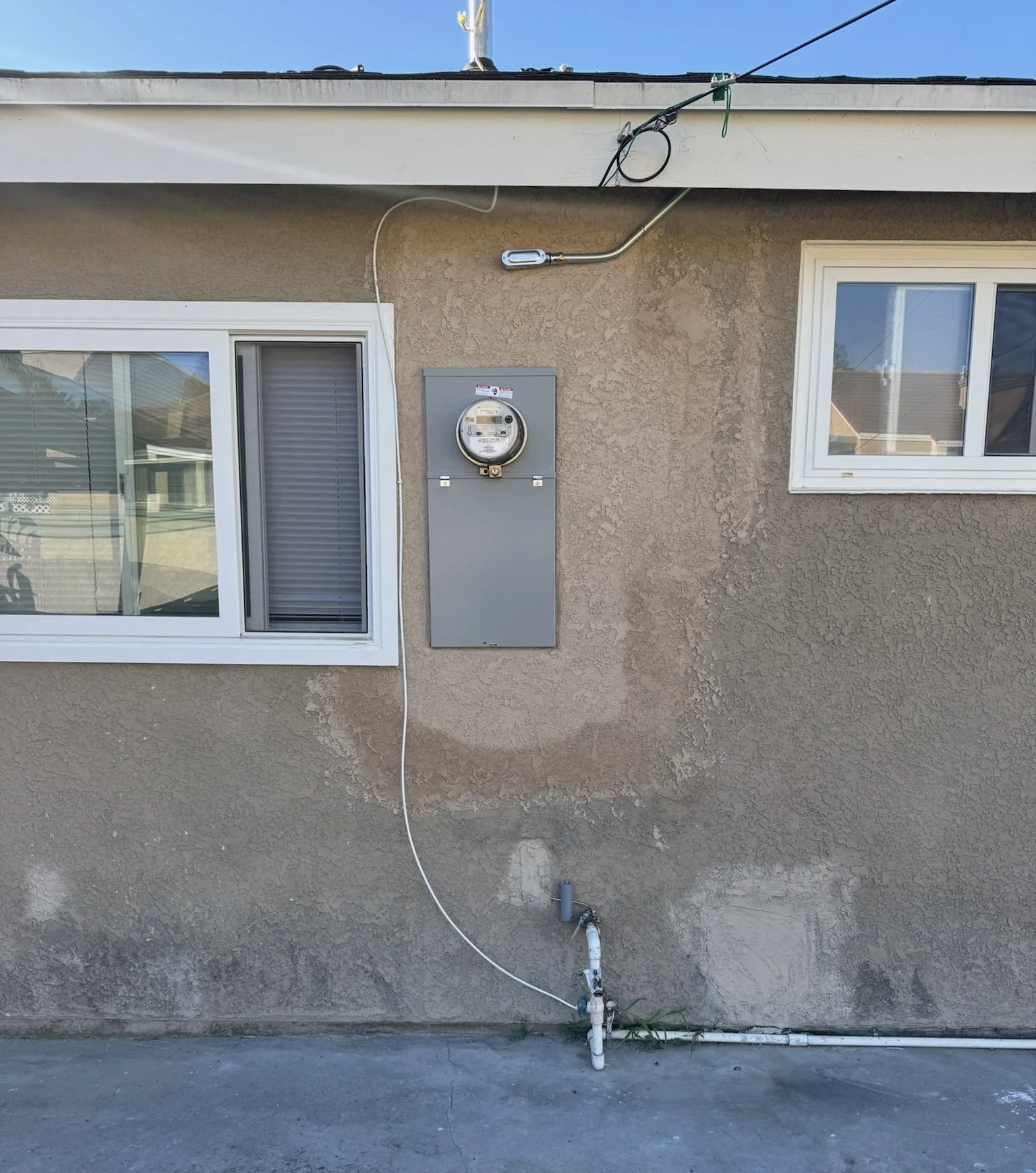 Exterior wall of a house with two windows, an electric meter box, and various cables and pipes. One window has blinds, and the other is clear. A cable runs from the roof to the meter, and pipes are connected to the ground.