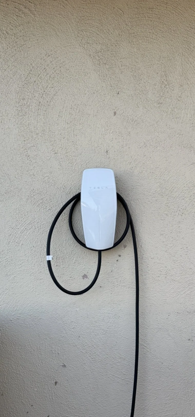 Tesla electric vehicle charger mounted on a beige textured wall with black charging cable hanging down.