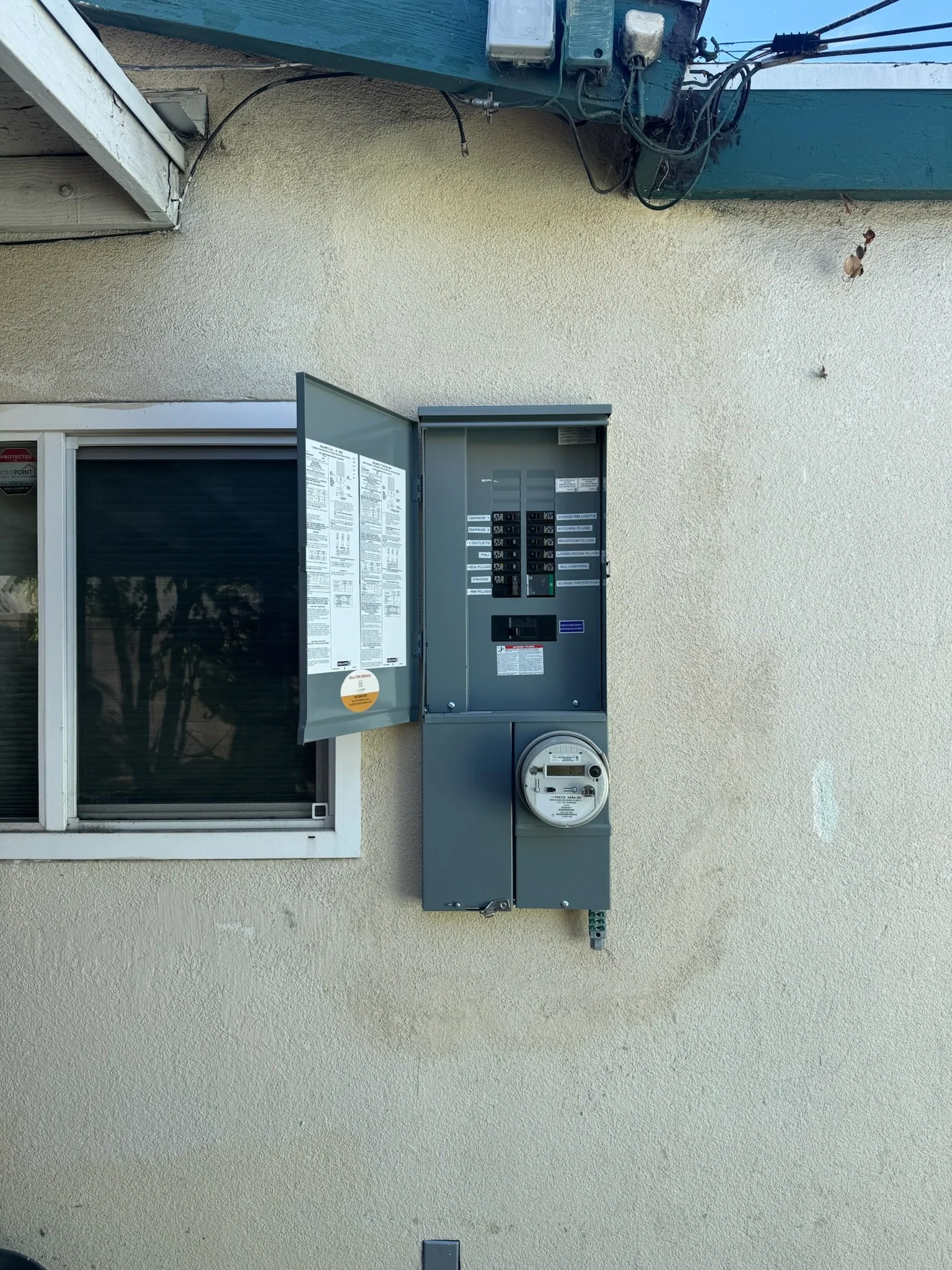 An electric meter and circuit breaker panel mounted on an exterior beige stucco wall next to a window with open blinds.