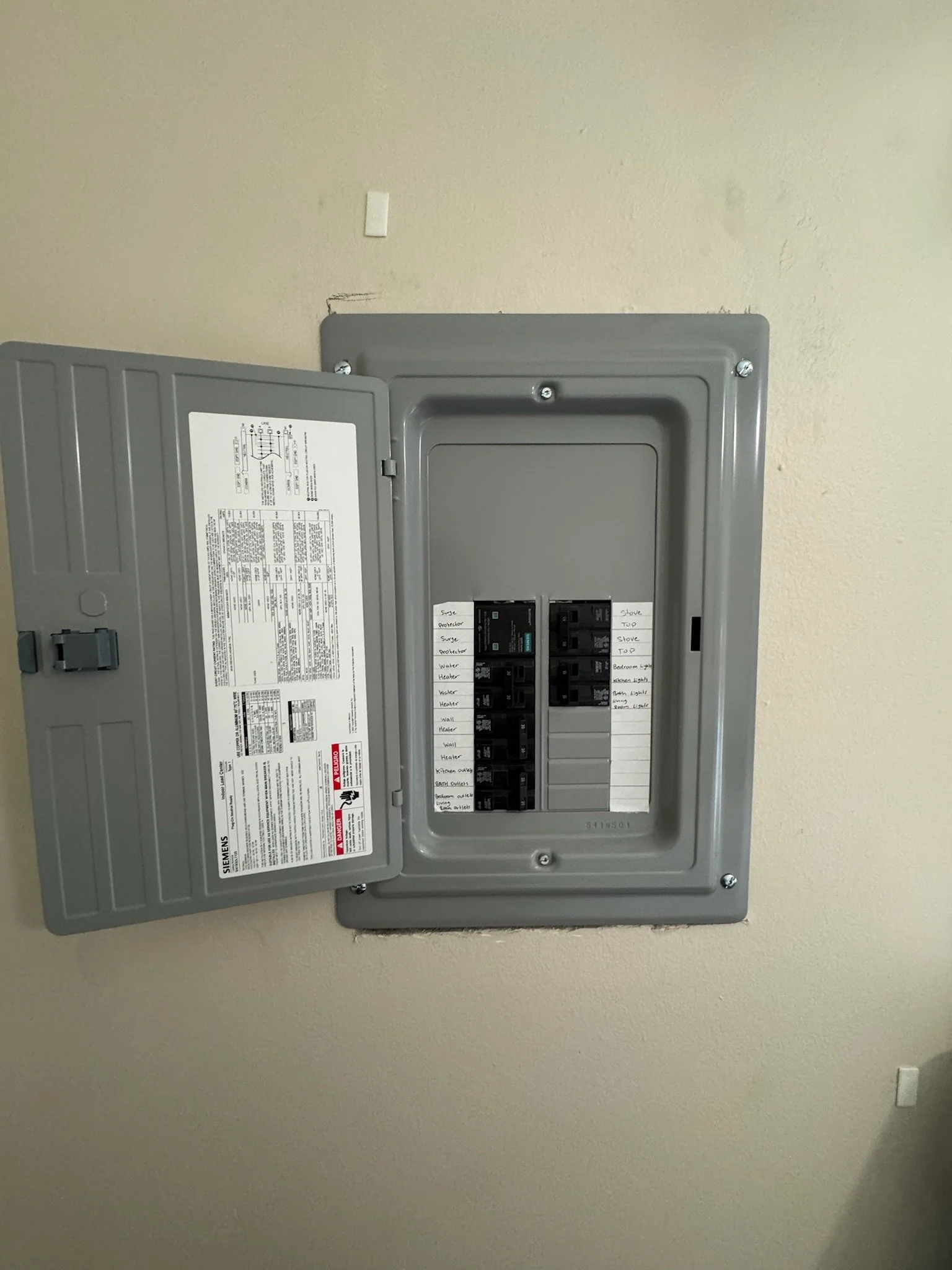 Open electrical breaker box with various circuit breakers and labels on a beige wall.