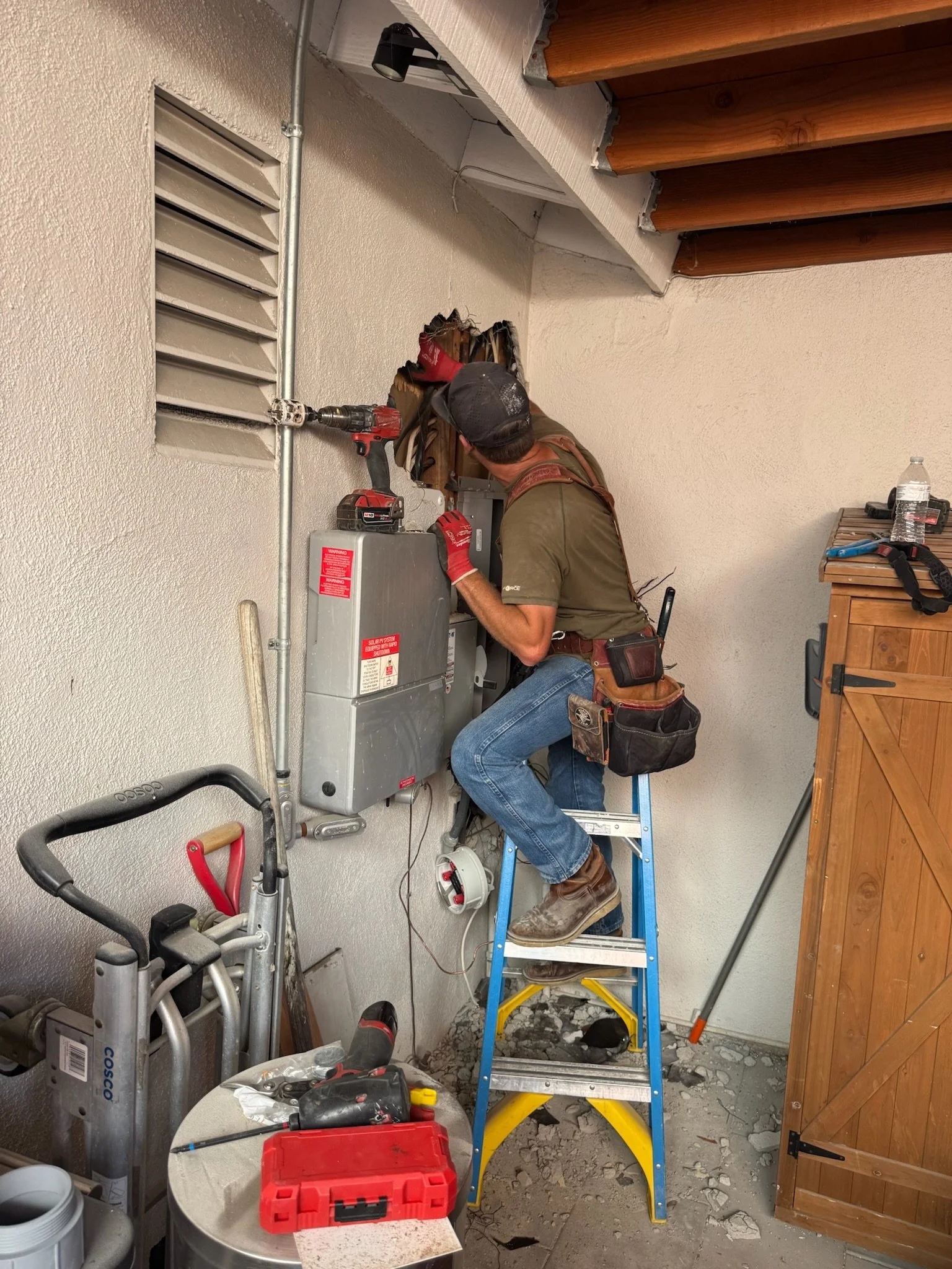 A man in a work uniform and safety gear, standing on a step ladder, repairing electrical or HVAC equipment on a wall in a maintenance or utility room.
