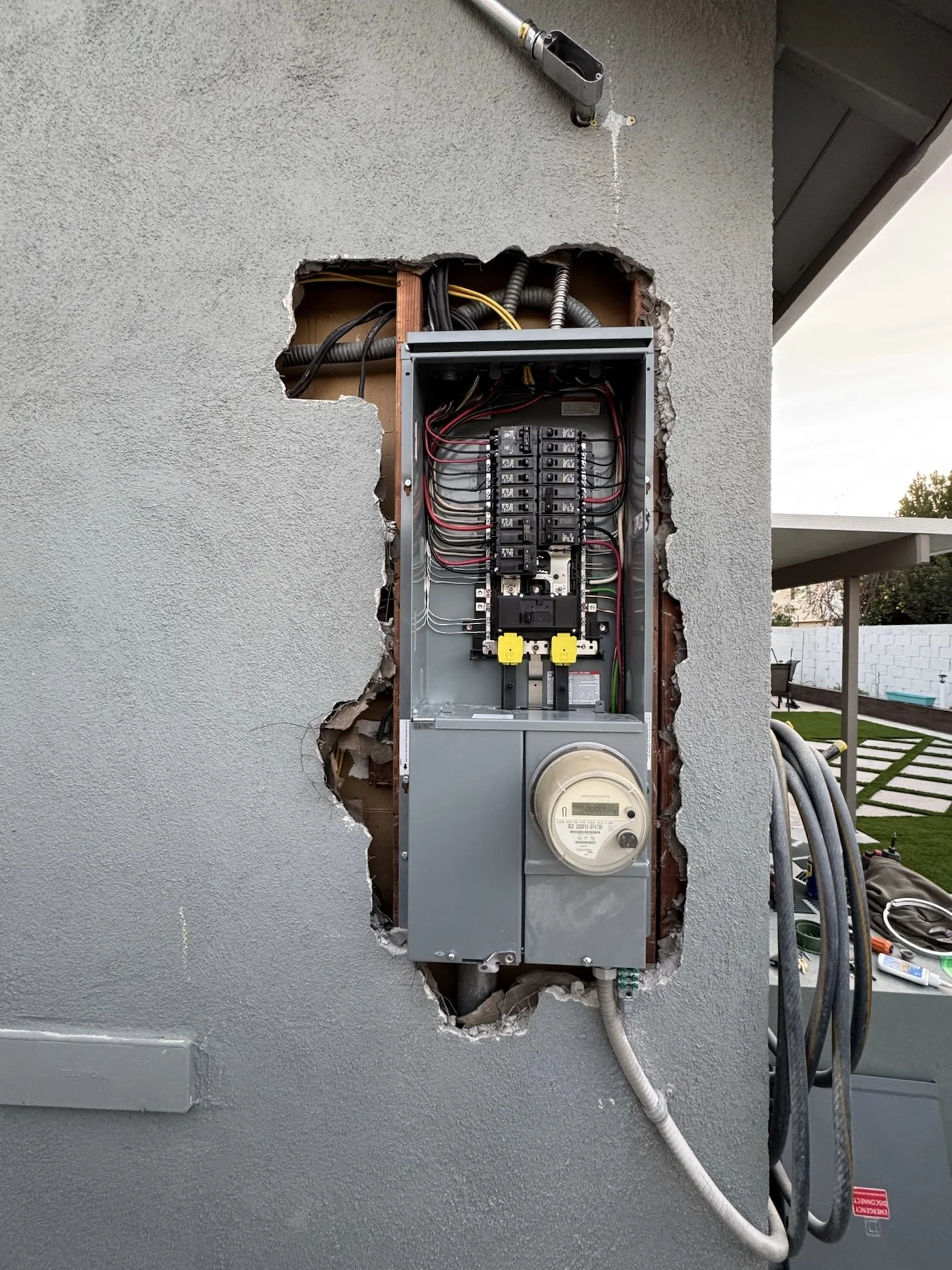 An open electrical meter box mounted on a gray wall with visible wiring and circuit breakers. A utility meter is attached to the bottom of the box, and there are hoses and tools on a table nearby, indicating ongoing electrical work.