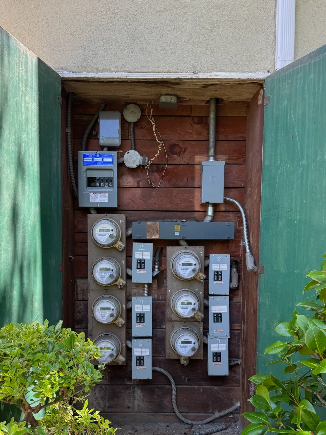 Outdoor electrical meters and panels installed within a small wooden enclosure, flanked by green foliage.