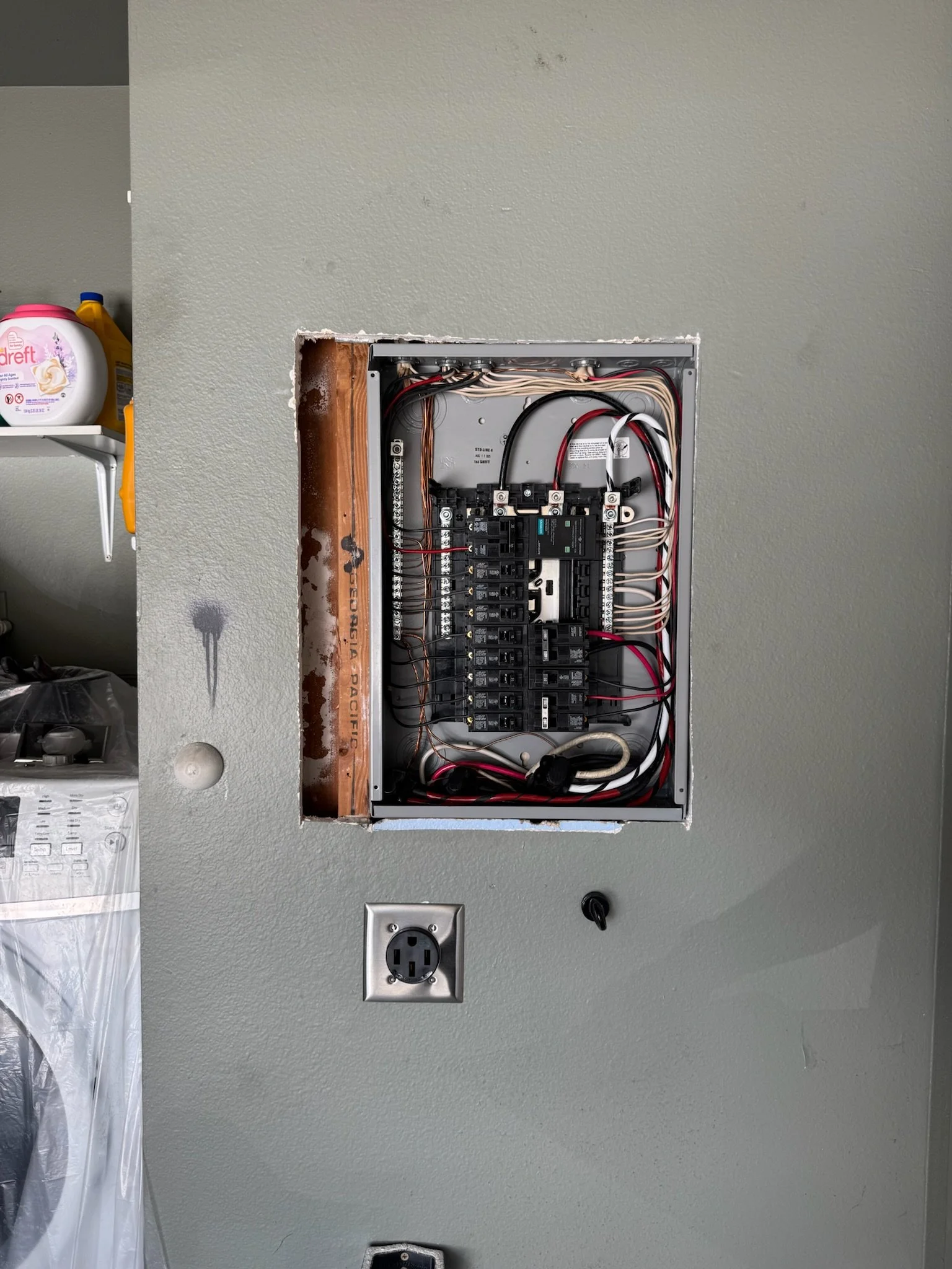 Open electrical panel in a wall with wiring and circuit breakers visible, next to a power outlet and a black knob on a gray wall.