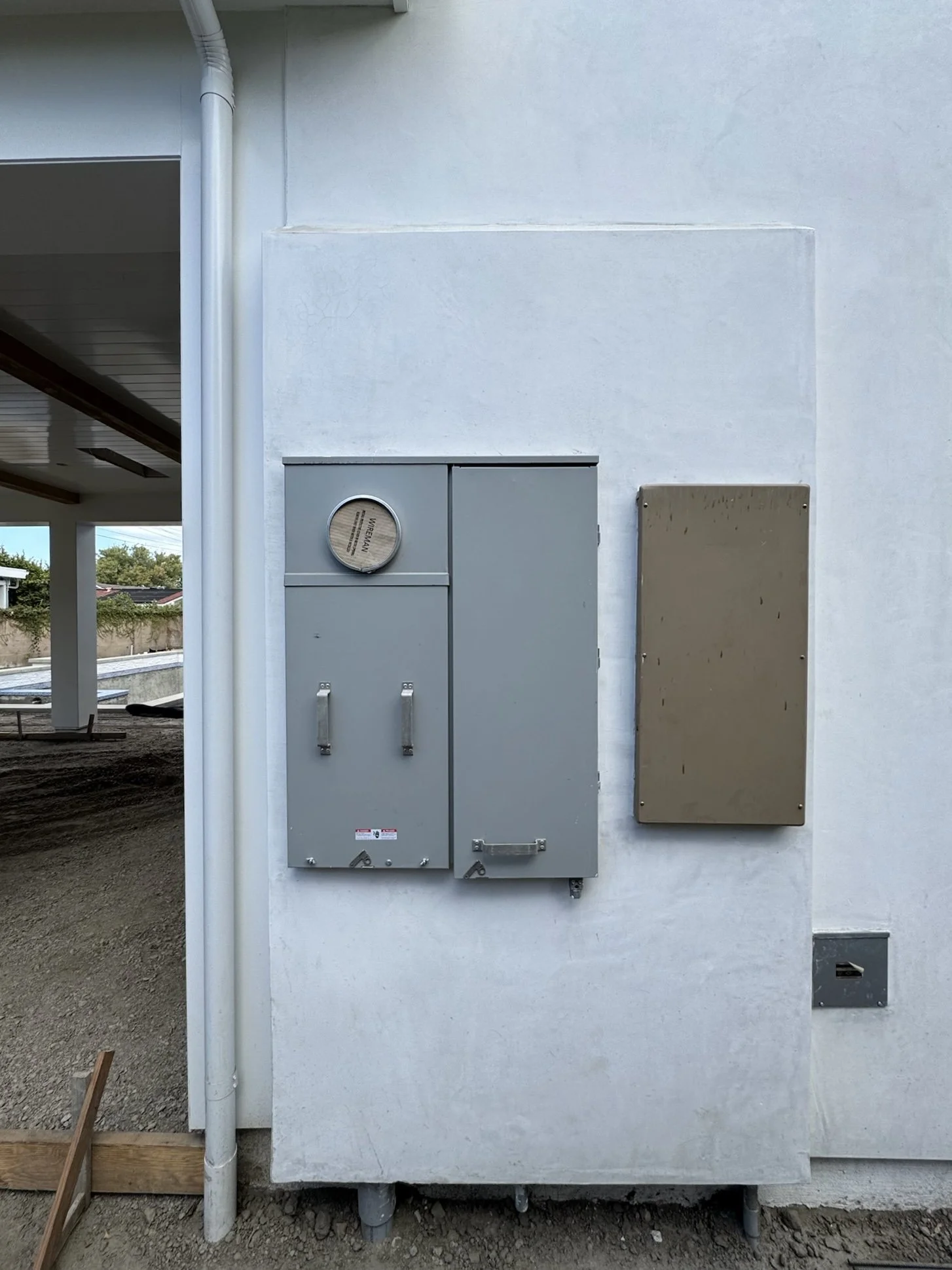 Outdoor view showing electrical meters and panels mounted on a white wall with a small patch of dirt and construction materials nearby.