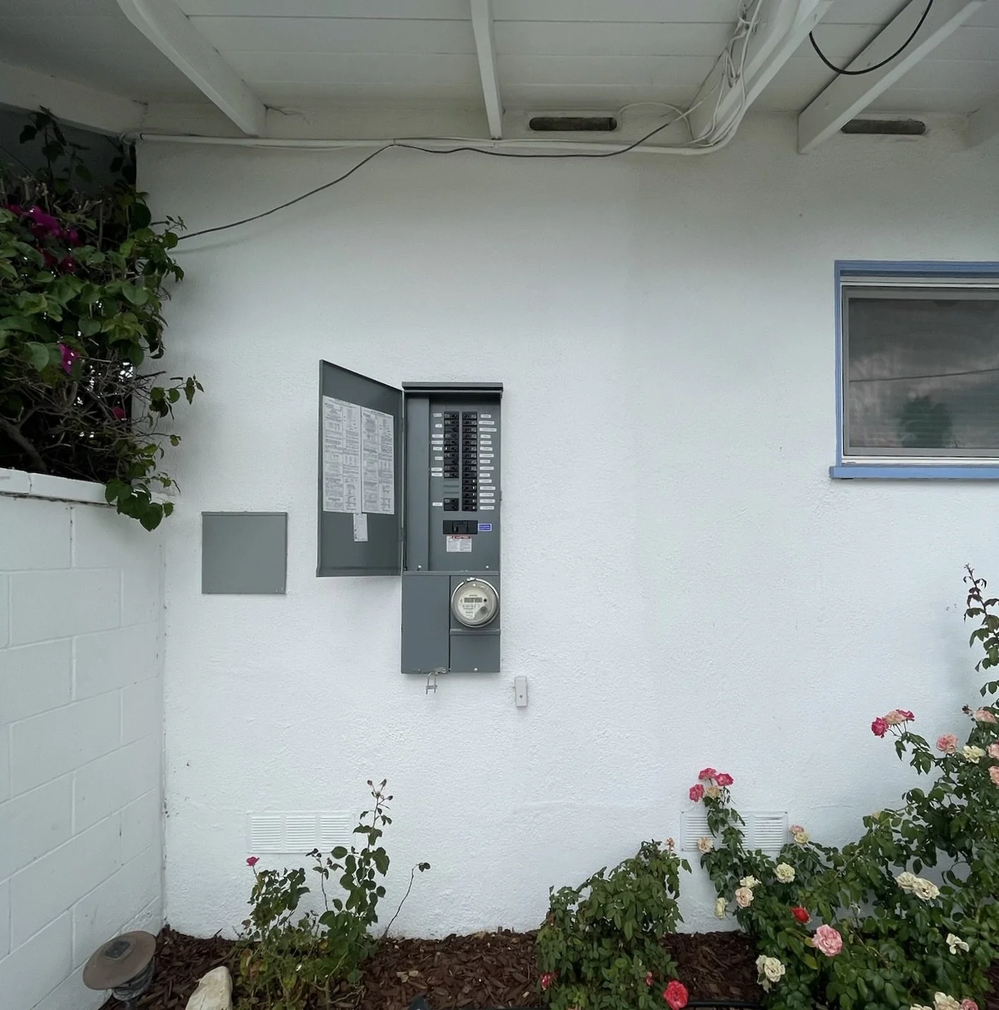 Exterior view of an electrical utility box on a white wall with plants and flowers below and a window to the right.