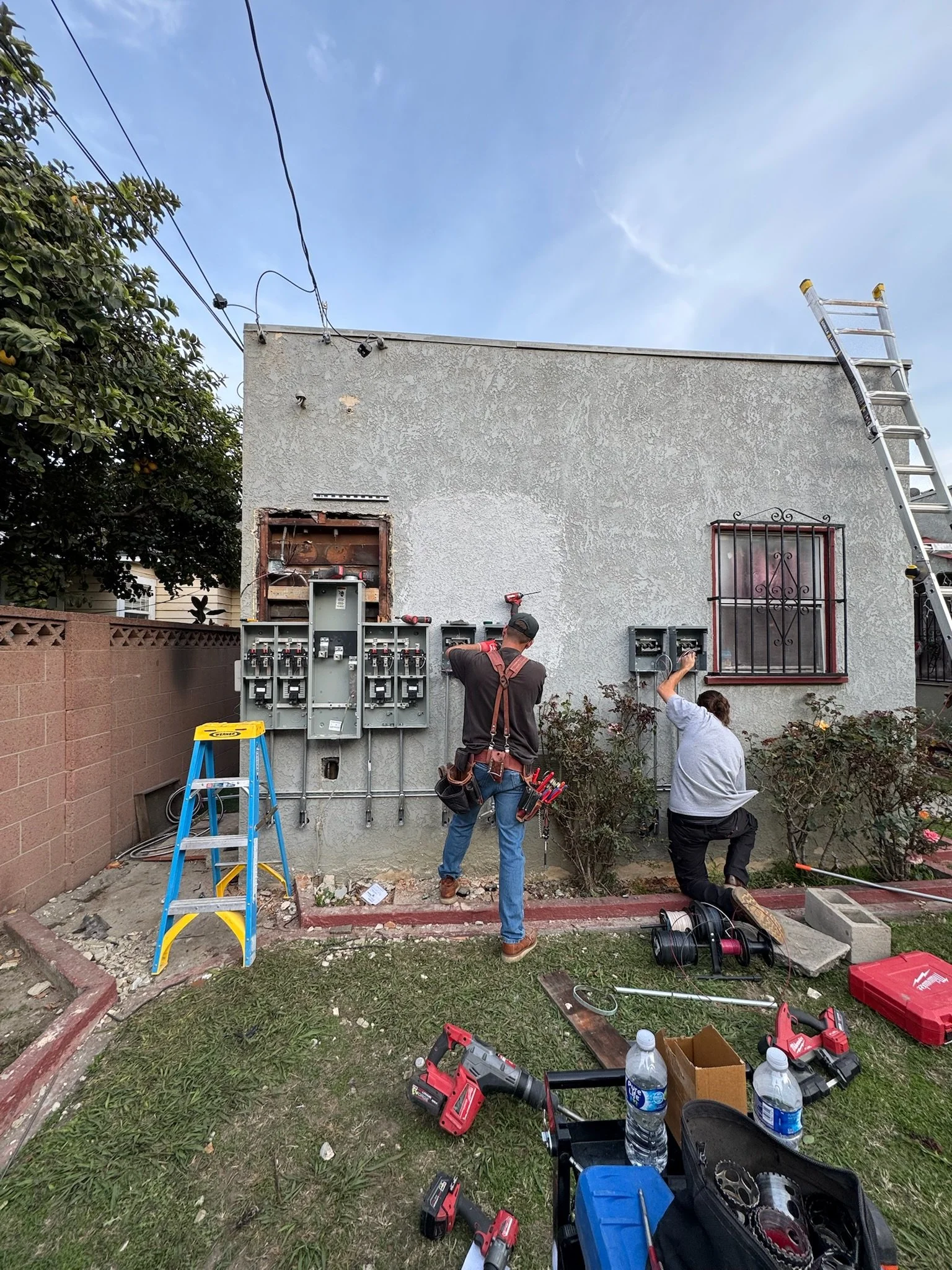Two workers repairing electrical meters on the exterior wall of a house with tools and equipment scattered on the ground.