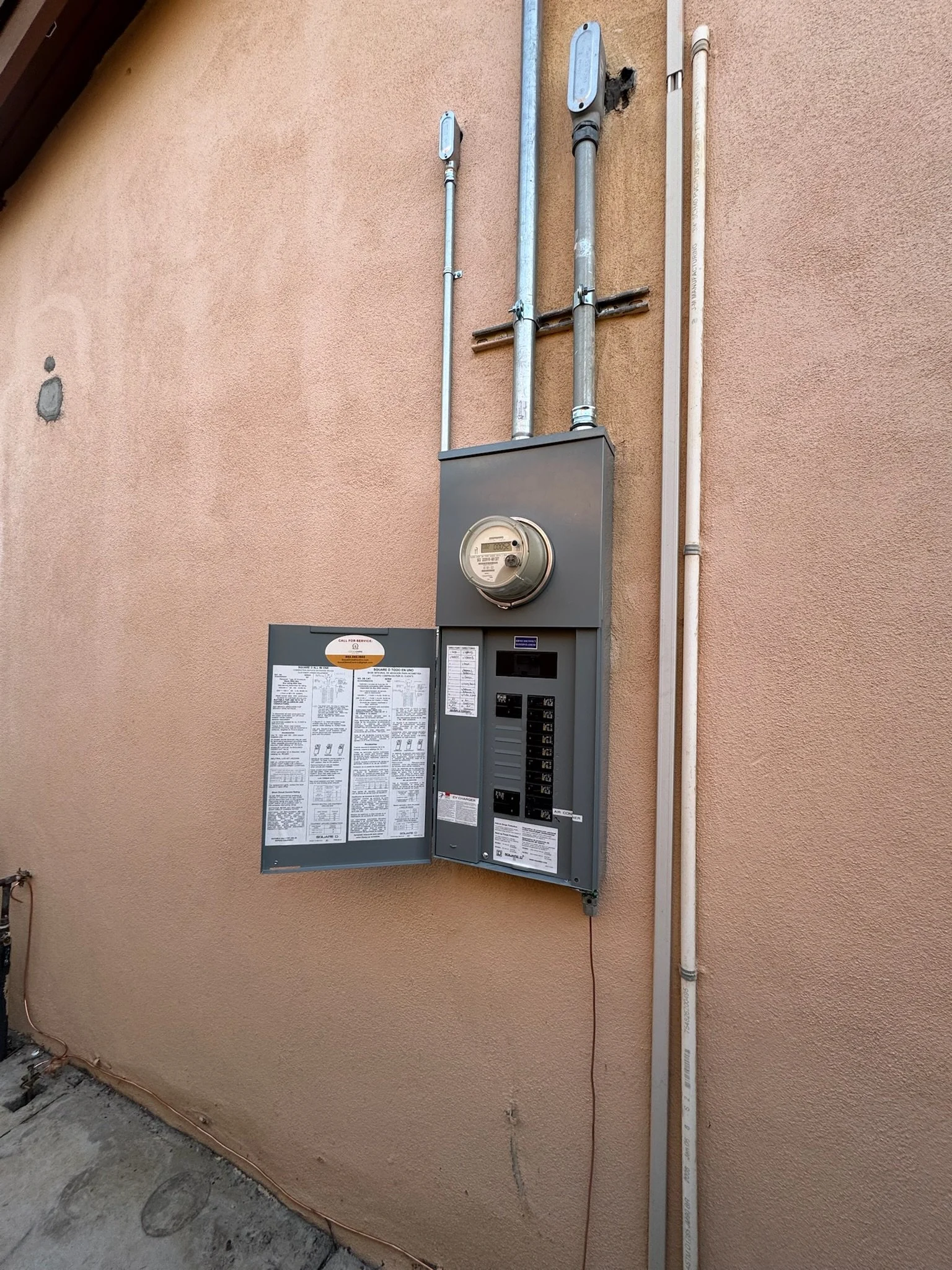 Electrical meter and breaker box mounted on a peach-colored exterior wall with metal conduit pipes running vertically above it.