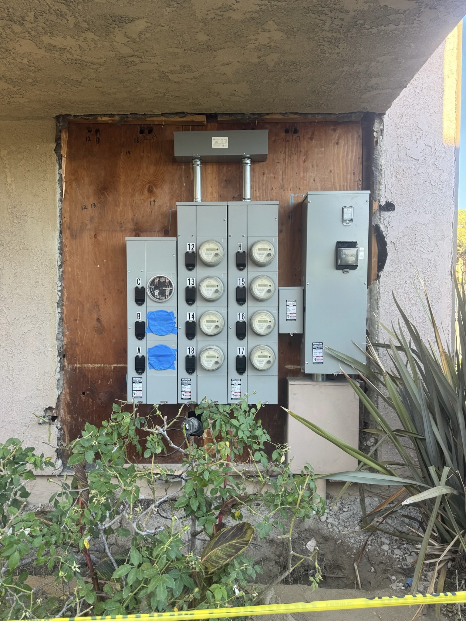 Exterior view of electrical meters and panels mounted on a wooden background with surrounding plants.