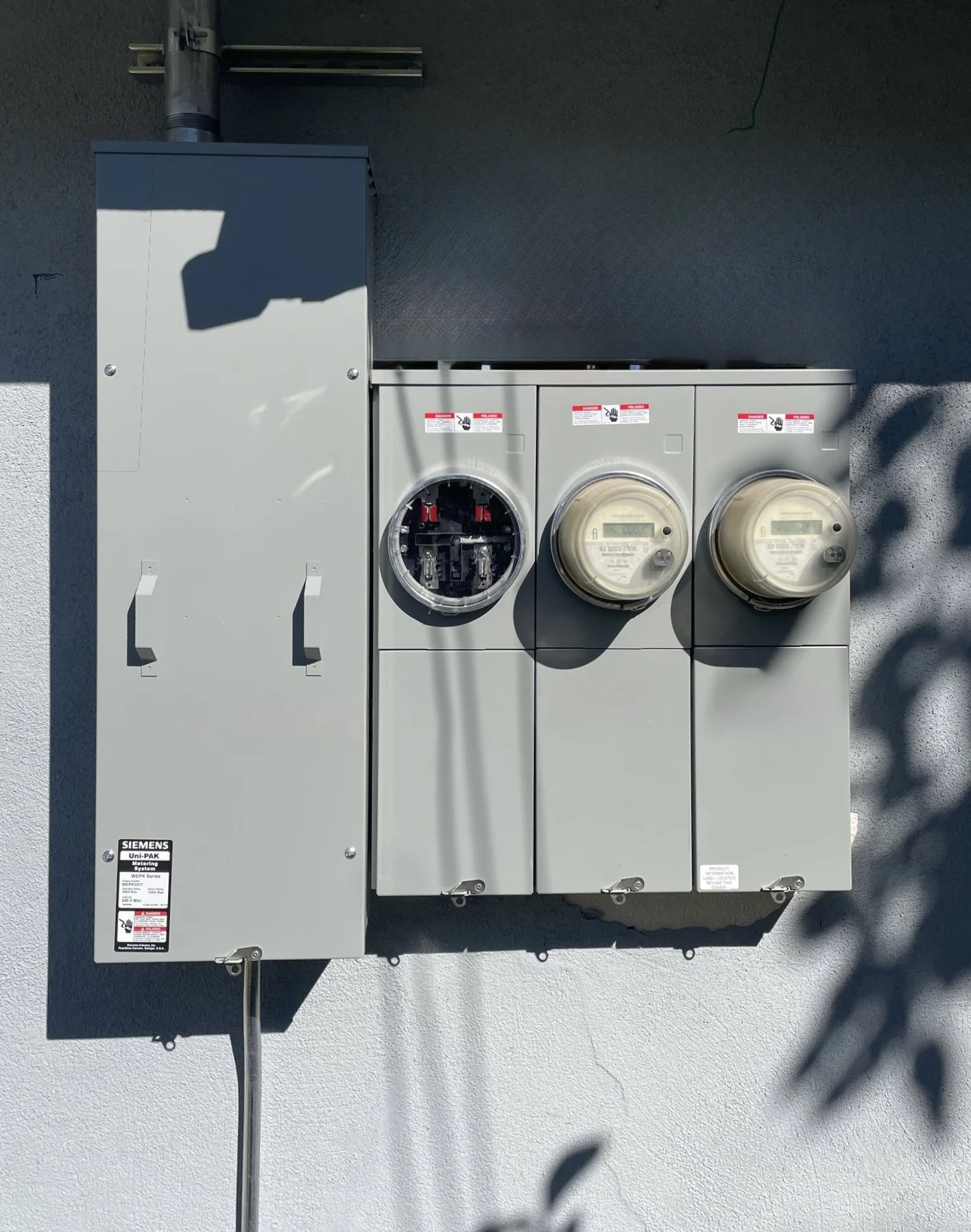 Electrical meters and a breaker box mounted on a wall outside, with shadows cast on the wall.