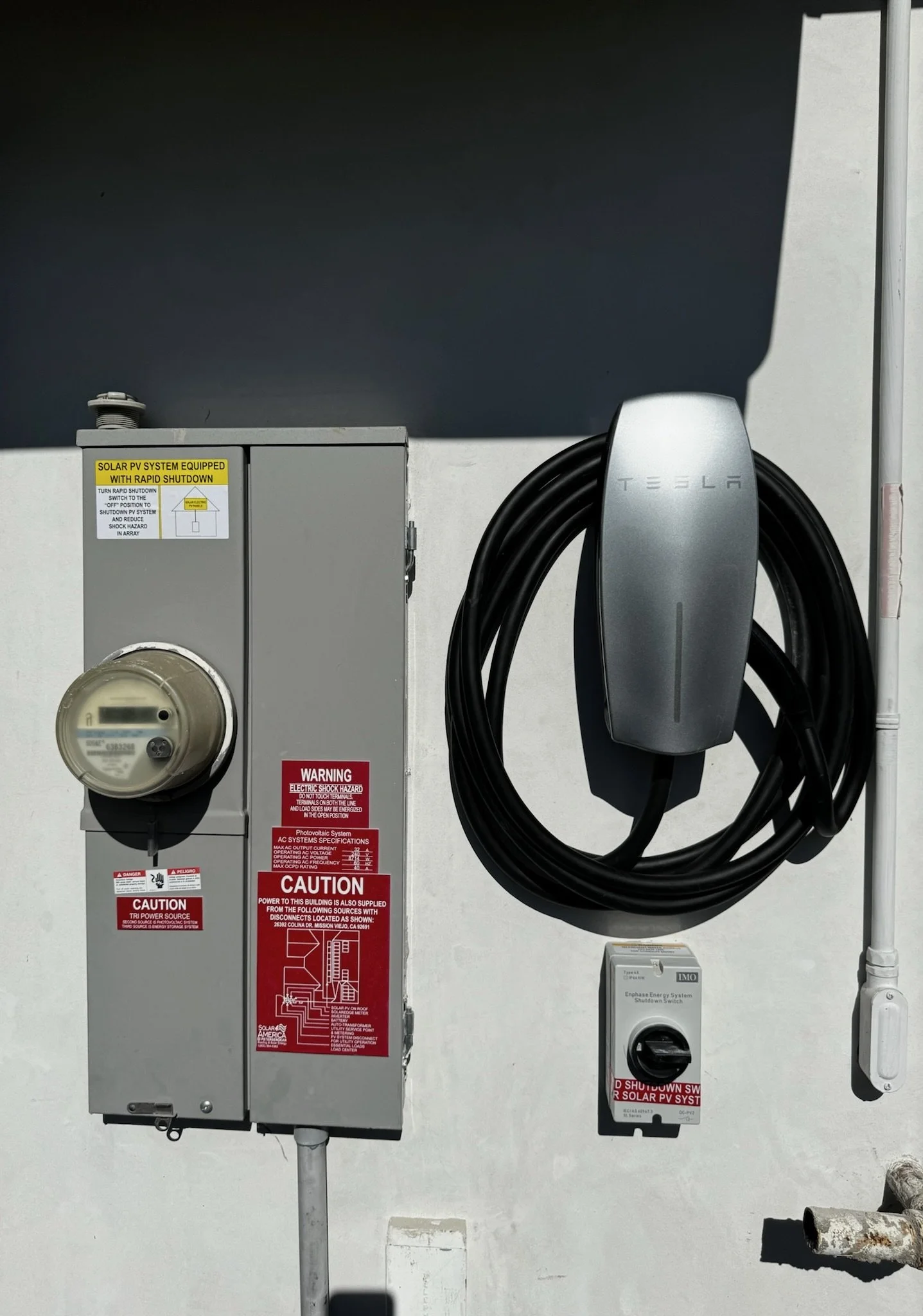 Electrical meter, Tesla charging station, and a disconnect switch on a white outdoor wall.