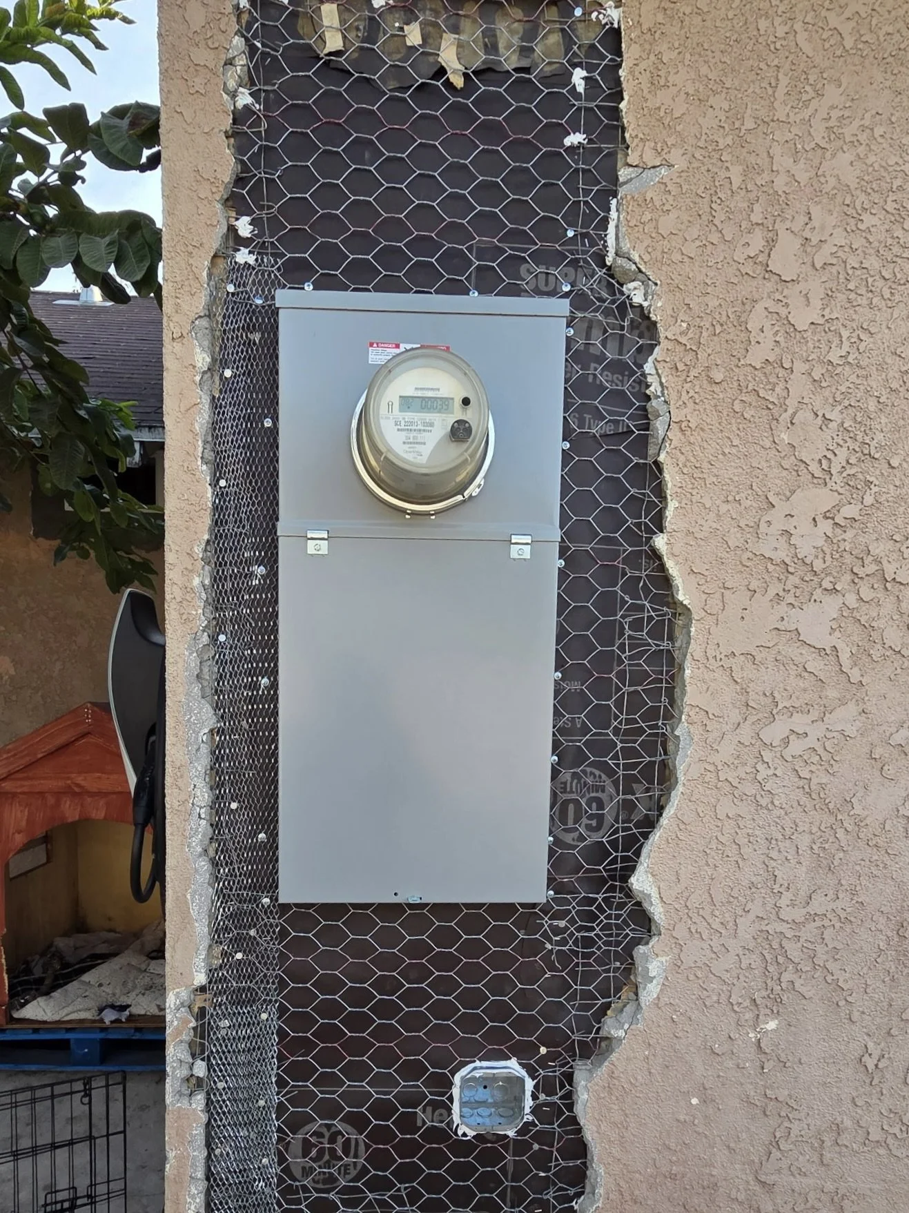 An outdoor electrical meter box mounted on a wall with exposed insulation and wire mesh, surrounded by a beige stucco wall and some green foliage on the left side.