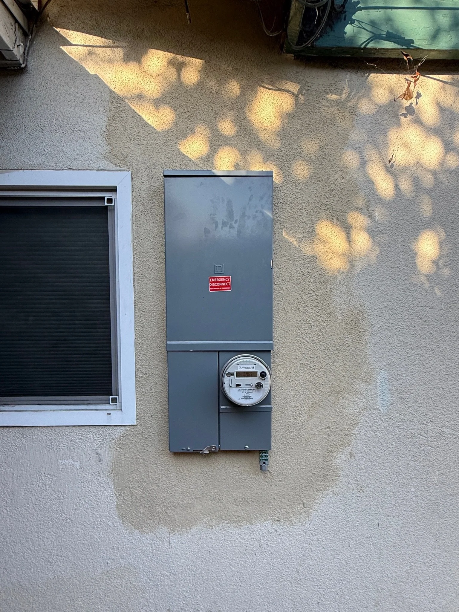 An electric meter and utility box mounted on a beige stucco wall outside a building, with shadows of tree leaves cast on the wall.