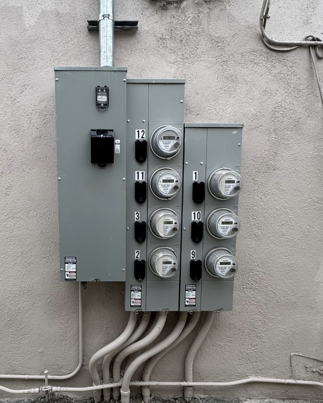 An outdoor electrical meter panel with multiple meters and disconnect switches mounted on a textured beige wall.