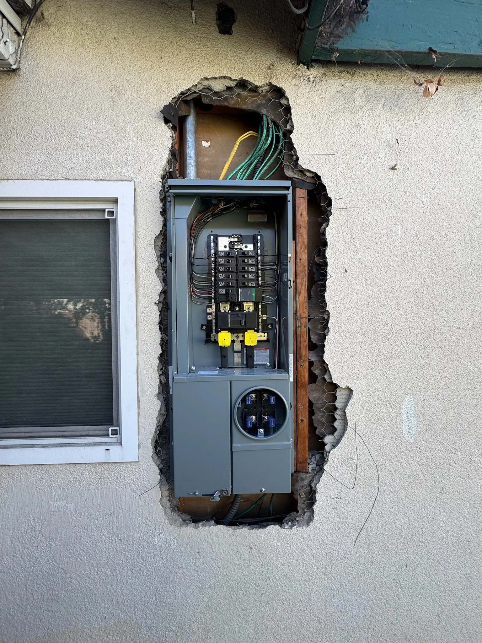 An outdoor electrical panel with wires and circuits, partially recessed in a wall with a surrounding rough cut opening.