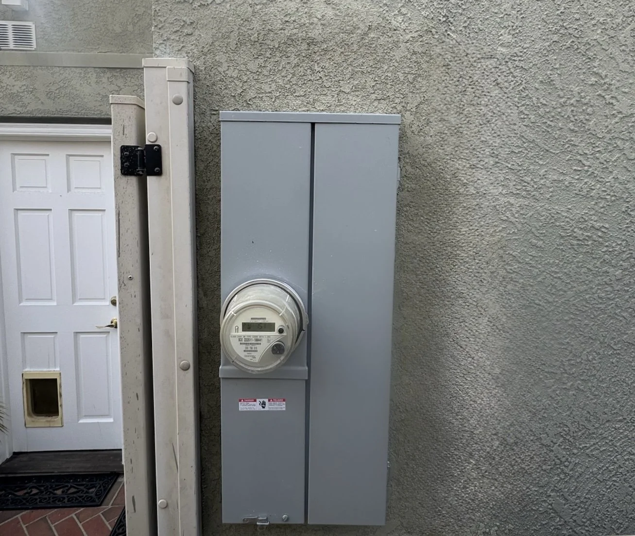 An outdoor electrical meter box installed on a beige stucco wall next to a white door.