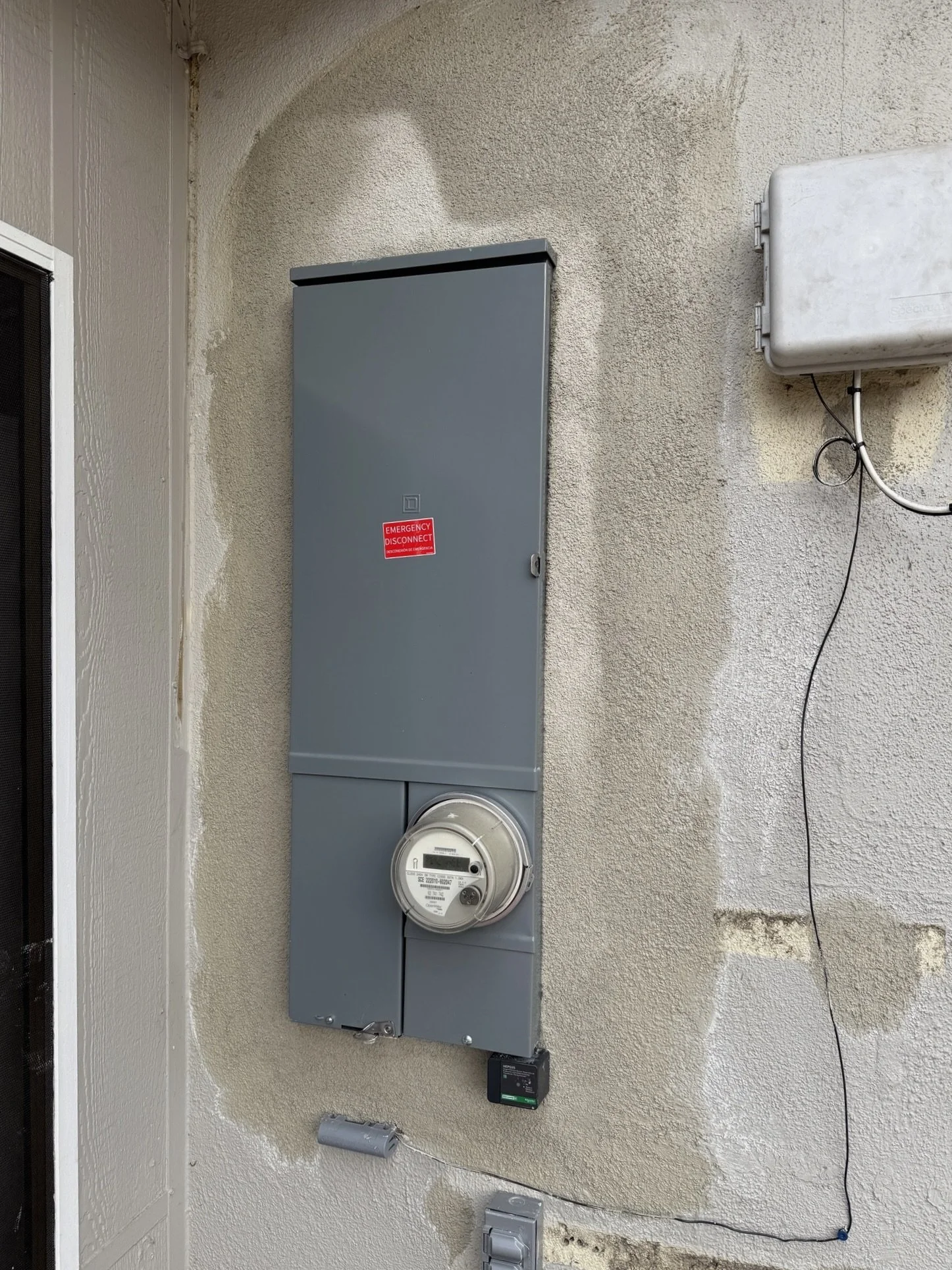 An outdoor electrical meter and panel mounted on a textured beige wall, with a cable hanging loosely and a smaller gray box nearby.