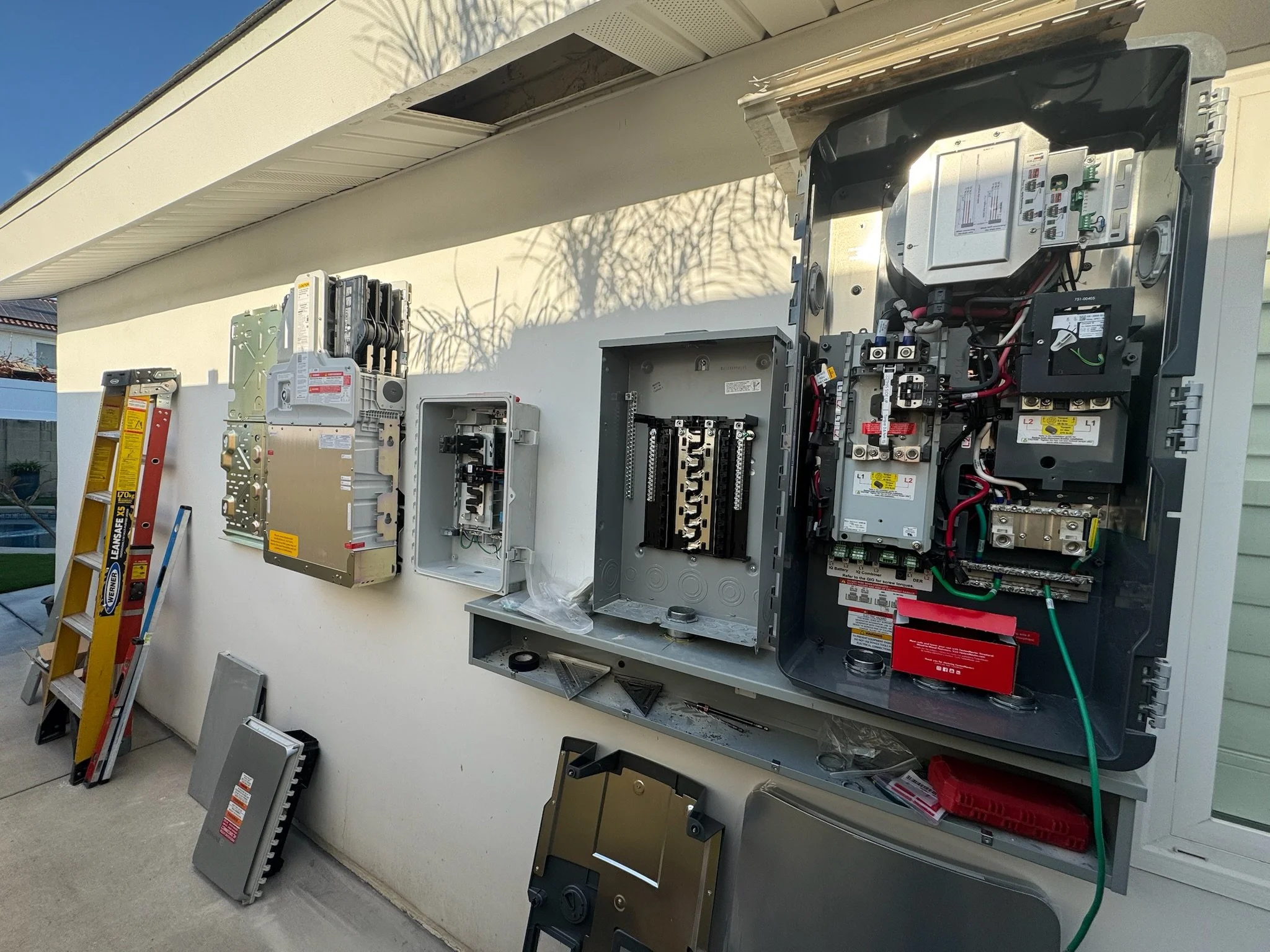 Outdoor view of an electrical panel and circuit breaker boxes with tools, ladder, and parts, during installation or repair.