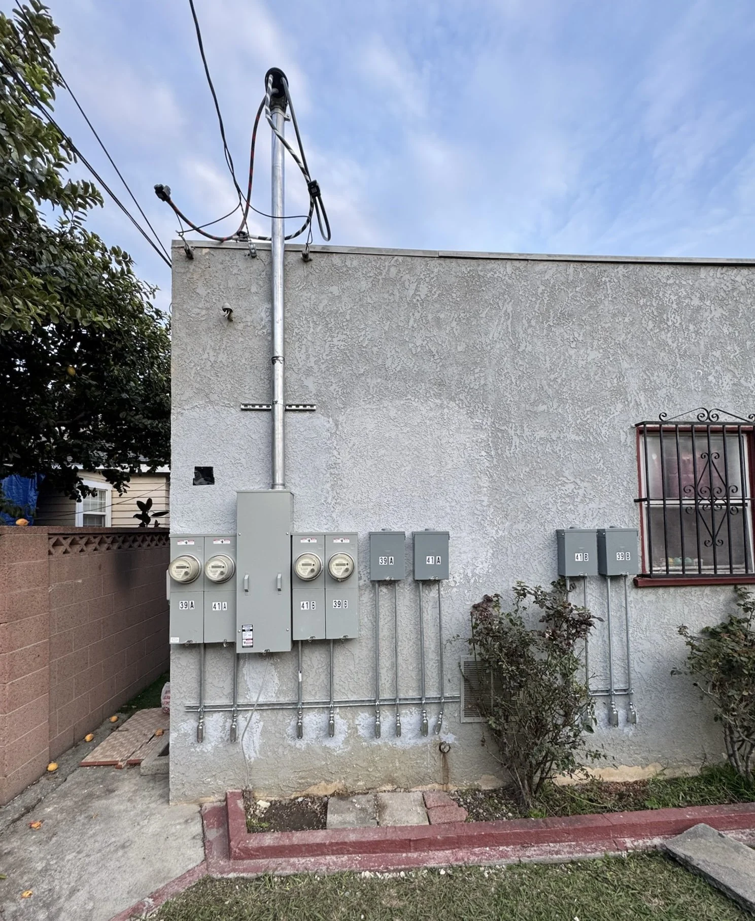 Electrical meters and boxes on a house wall with wiring and conduit, window with iron security bars, bushes, and a small garden bed in front.