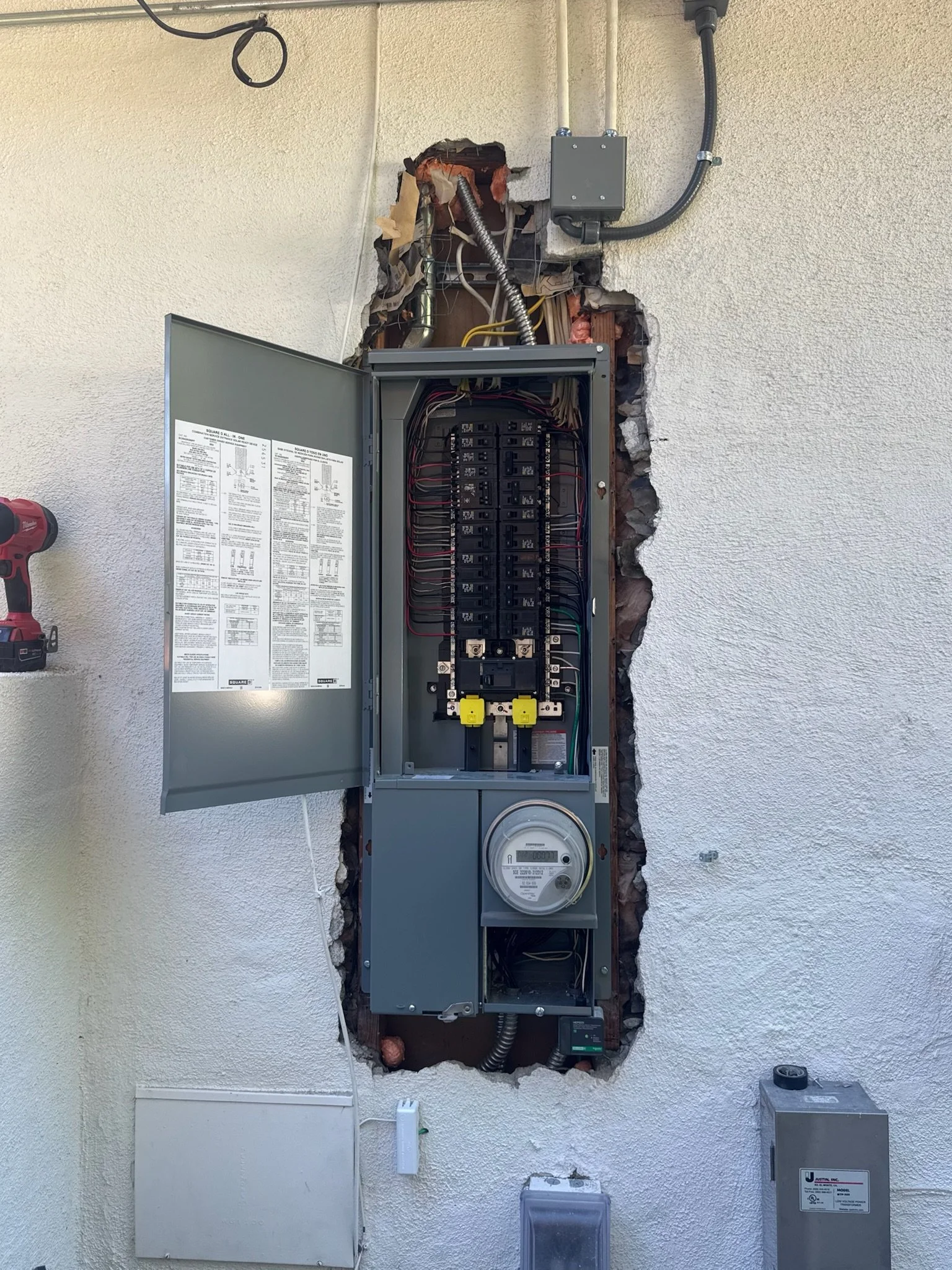 An open electrical breaker box installed in a wall with exposed wiring and circuit breakers, with a digital electricity meter at the bottom.