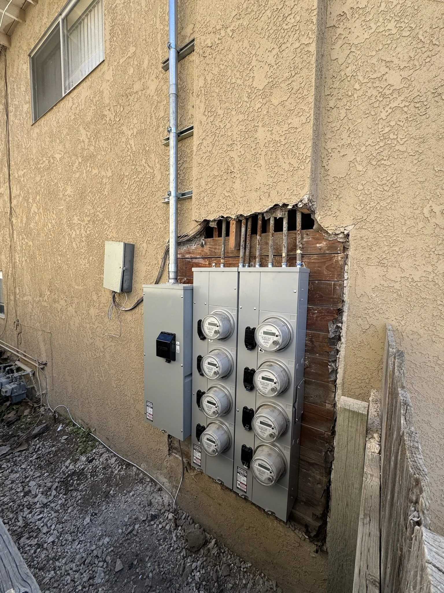 External electrical panel with multiple meters installed on the side of a beige stucco building, partially exposed wall showing wiring and wooden framing.