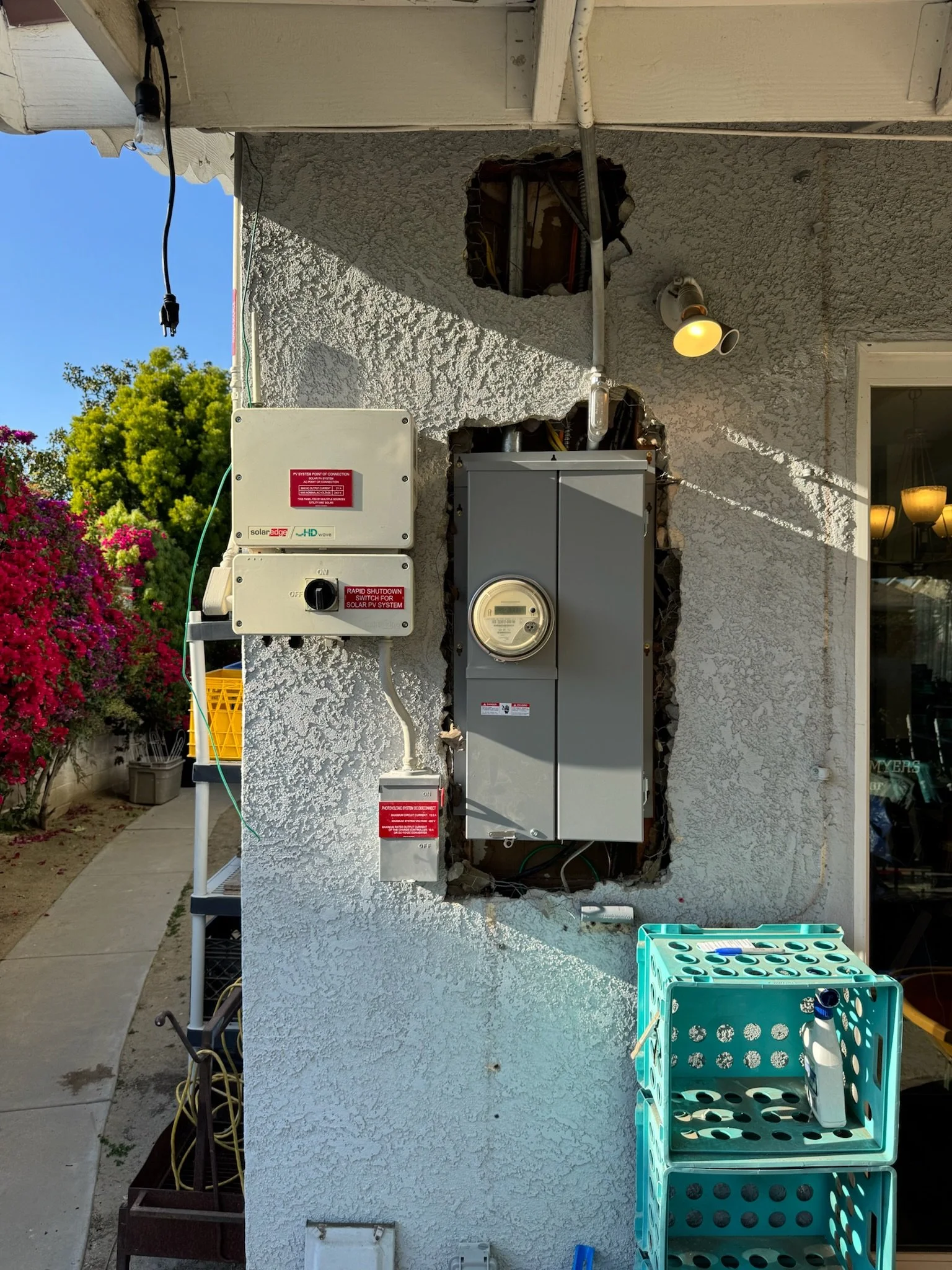 Outdoor electrical panel installation on a textured white wall with visible holes around the panel. A utility meter is mounted on the panel, and various wires are connected to it. There is a box labeled 'RAPID SHUTDOWN SWITCH FOR SOLAR PV SYSTEM,' an