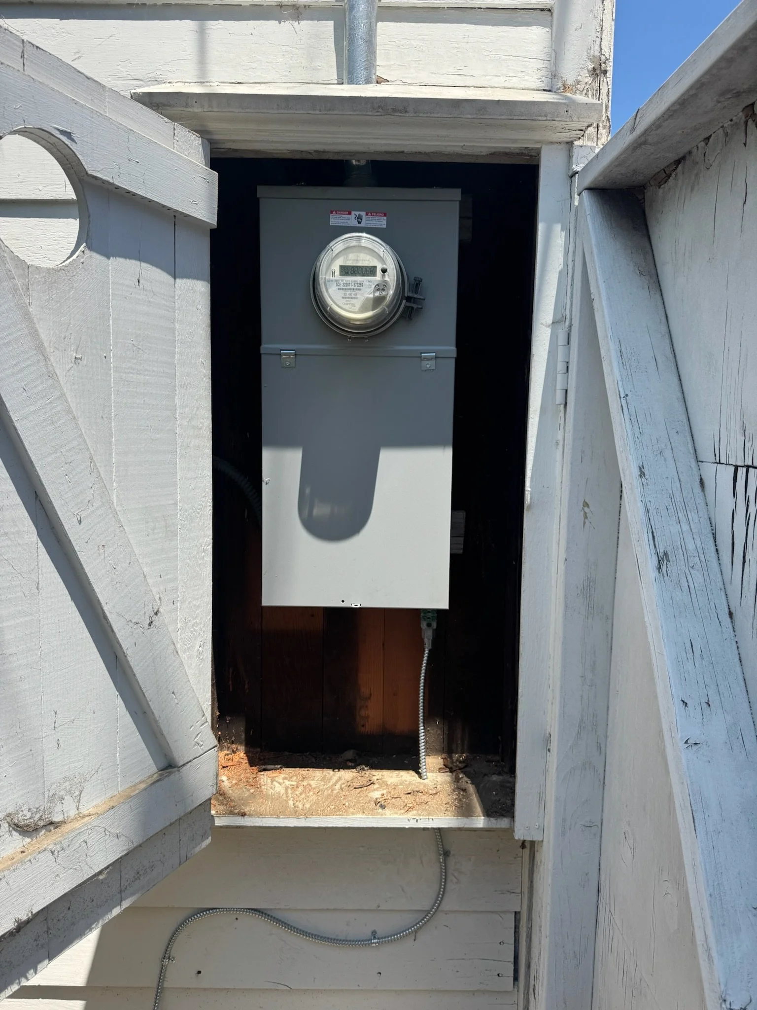 An electric meter attached to a gray box within a small wooden enclosure, with electrical conduits running into the box, against a background of light-colored wooden structures and clear blue sky.