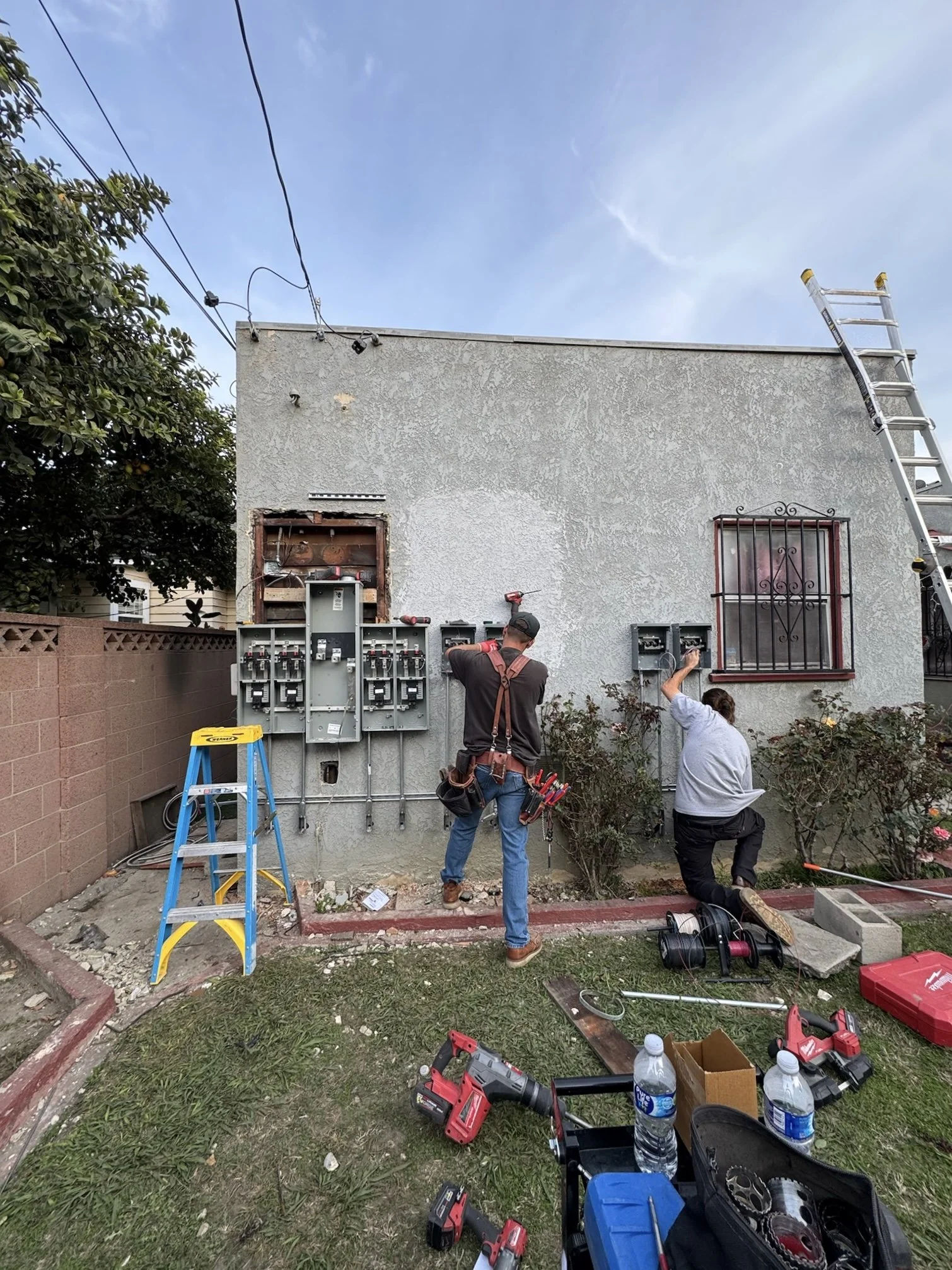 Two workers installing electrical panels on the exterior wall of a building, with a ladder leaning against the wall, tools and equipment on the ground, and a ladder on the right side of the building.