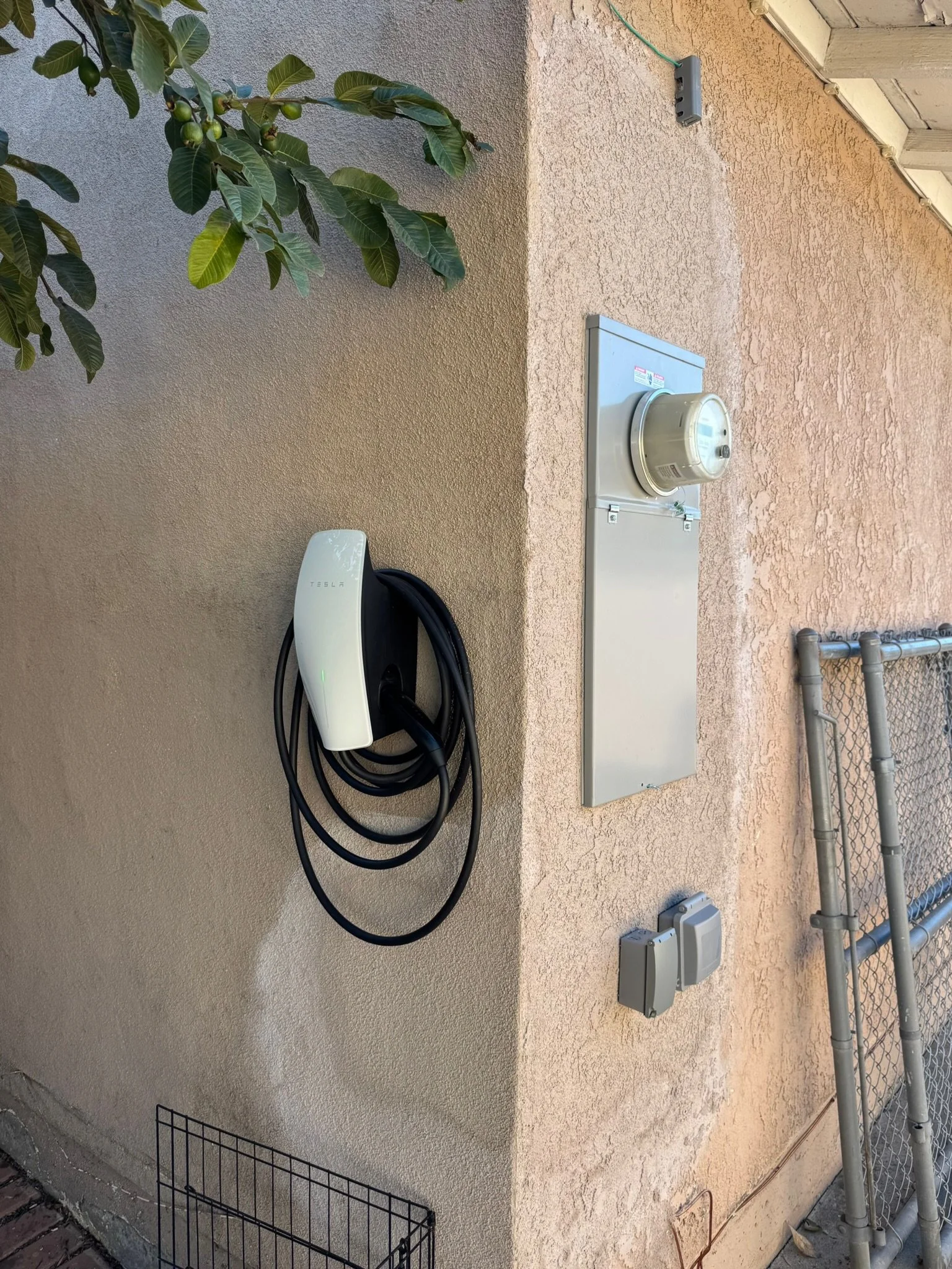 Electric vehicle charging station with a Tesla charger, electrical boxes, and a metal fence in front of a beige stucco wall.