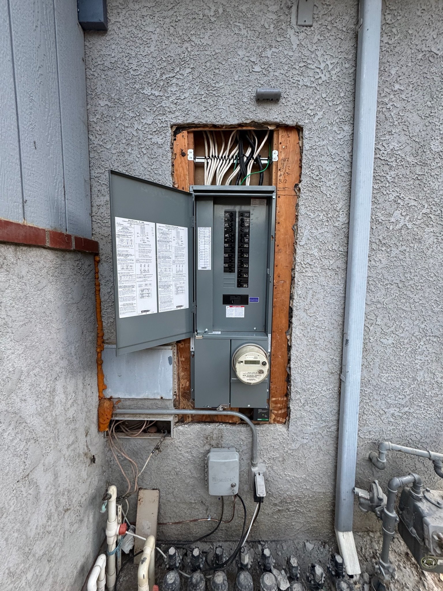 An electrical meter, circuit breaker panel, and wiring near a concrete wall.