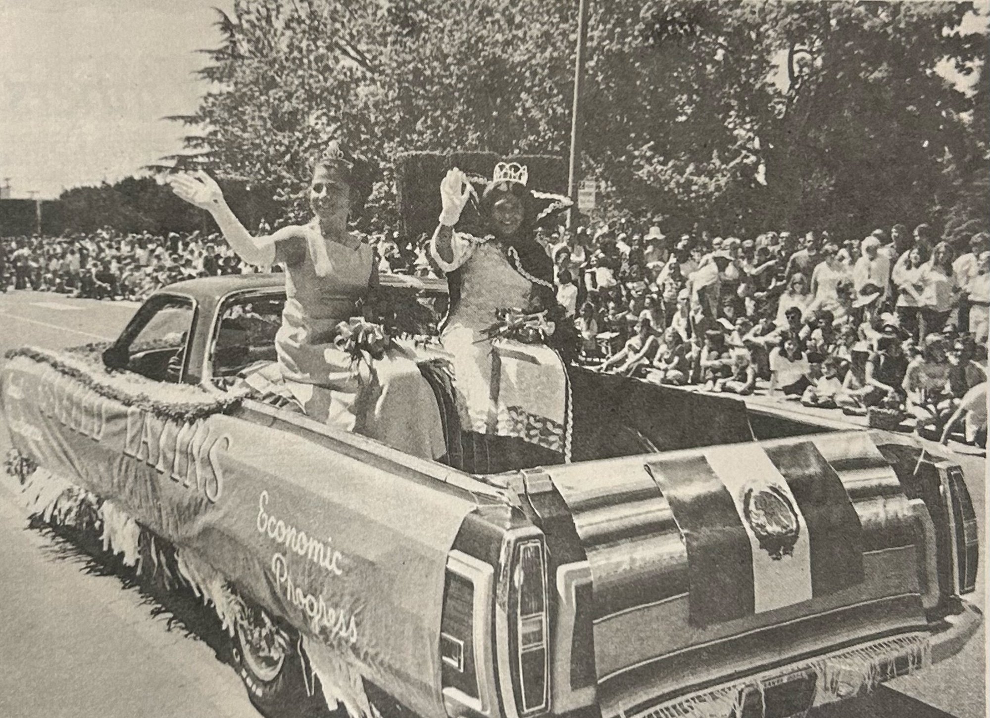 Black and white photo of a parade with a decorated vehicle carrying people waving