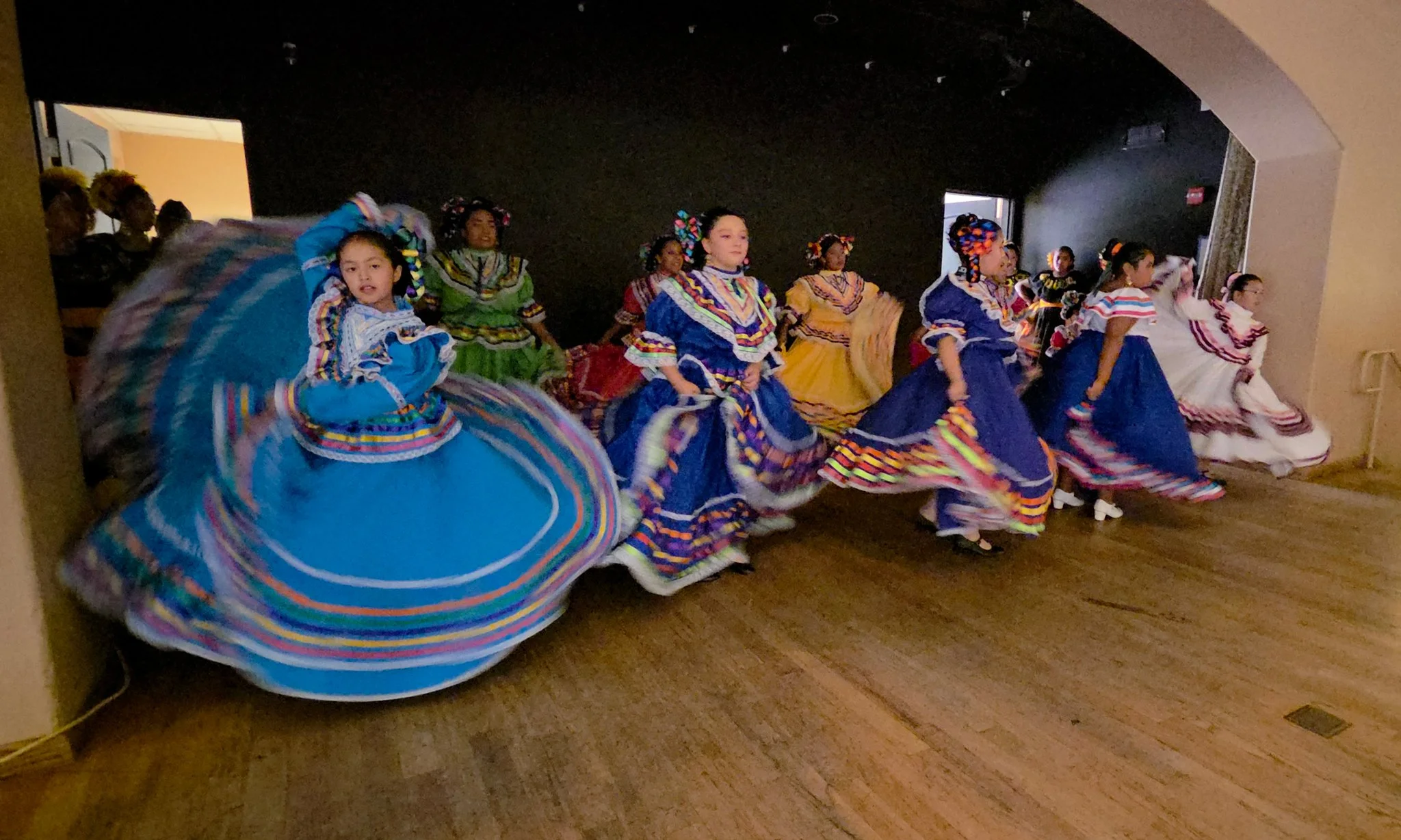Young girls dressed in colorful traditional Mexican dresses with floral headpieces performing a dance indoors.