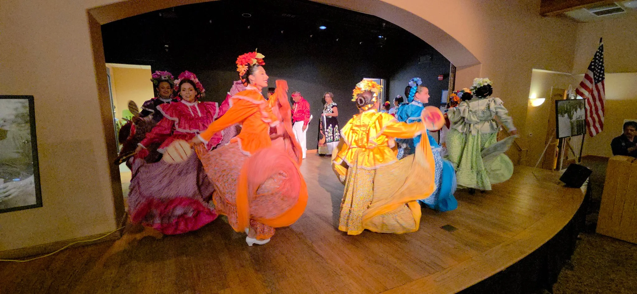 Group of women dressed in colorful, traditional Mexican dresses performing a folk dance on stage.
