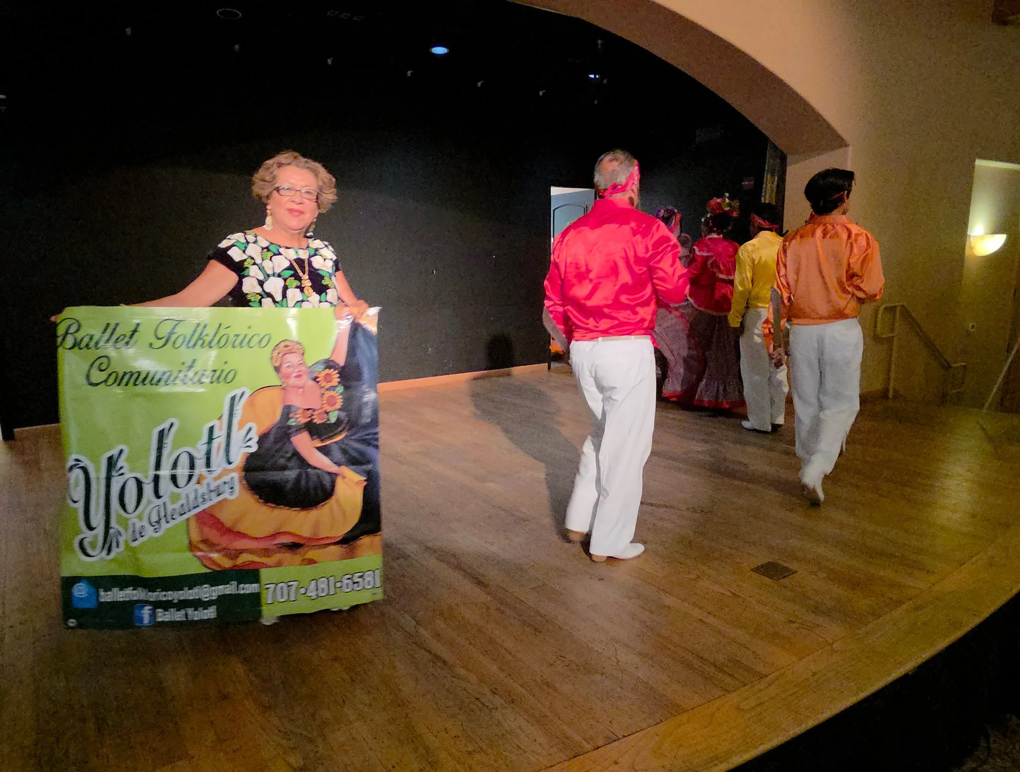 A woman holding a green promotional sign for a ballet folklórico community event, with people dressed in colorful clothing in the background on a stage.