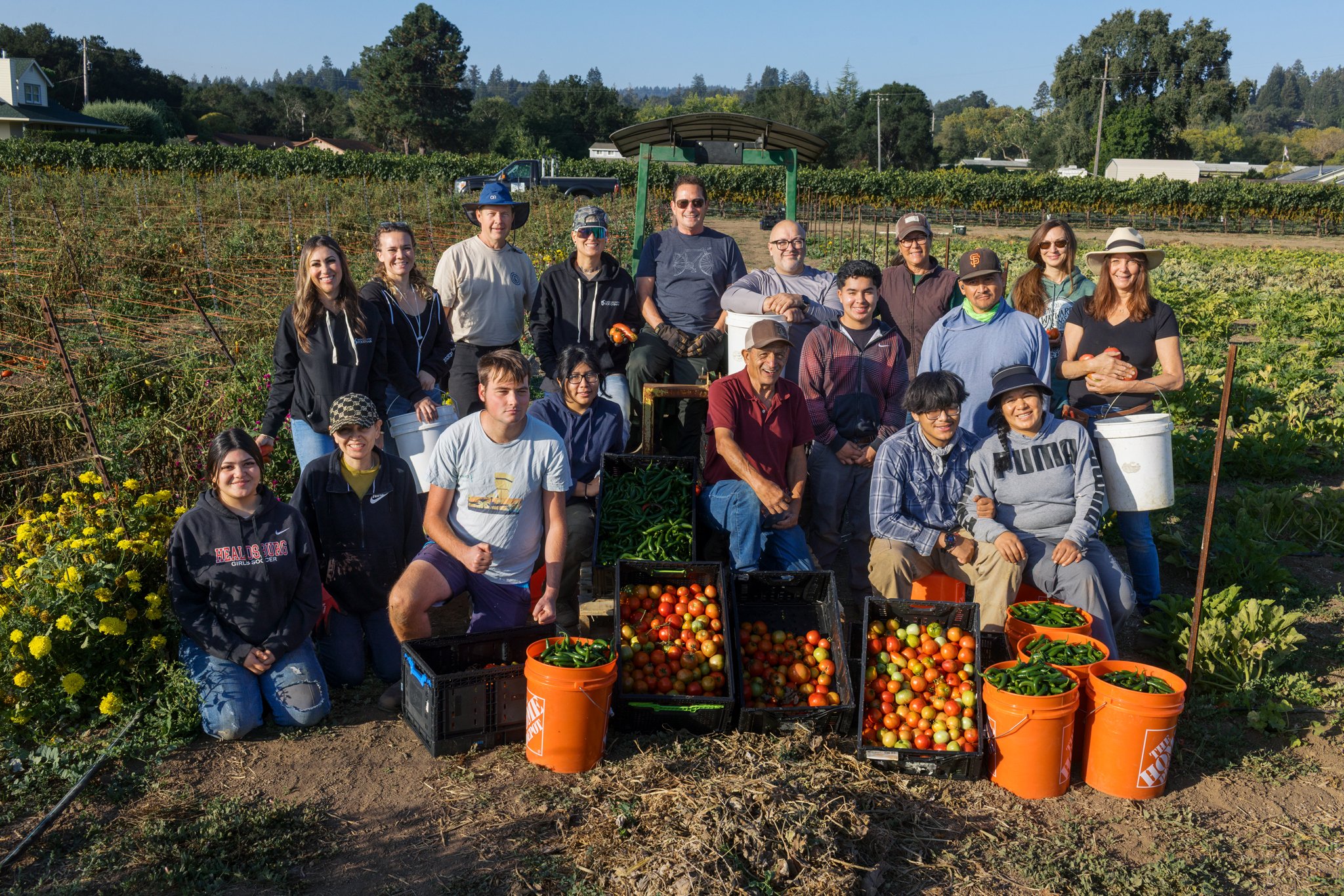 Group of people at a farm with baskets of tomatoes and peppers, surrounded by rows of crops and trees in the background.