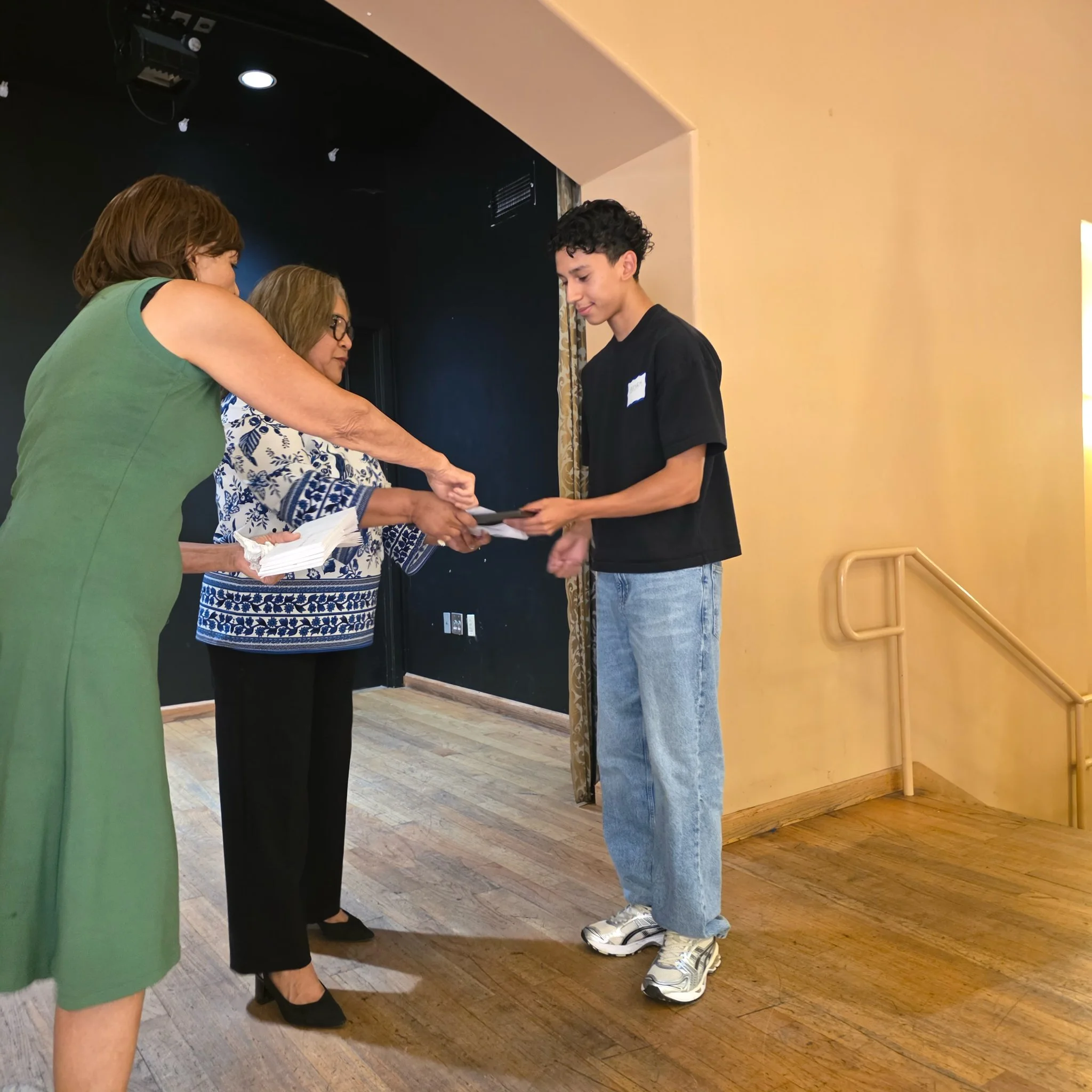Two women and a young man with curly hair are standing in a room with wooden flooring. The women are engaged in a conversation with the young man, who is holding a picture frame. One woman has glasses and is wearing a blue and white patterned top, while the other woman is in a green top. The man is dressed in a black shirt and jeans, with sneakers on the floor. The scene appears to be in an indoor setting with plain walls and minimal decor.