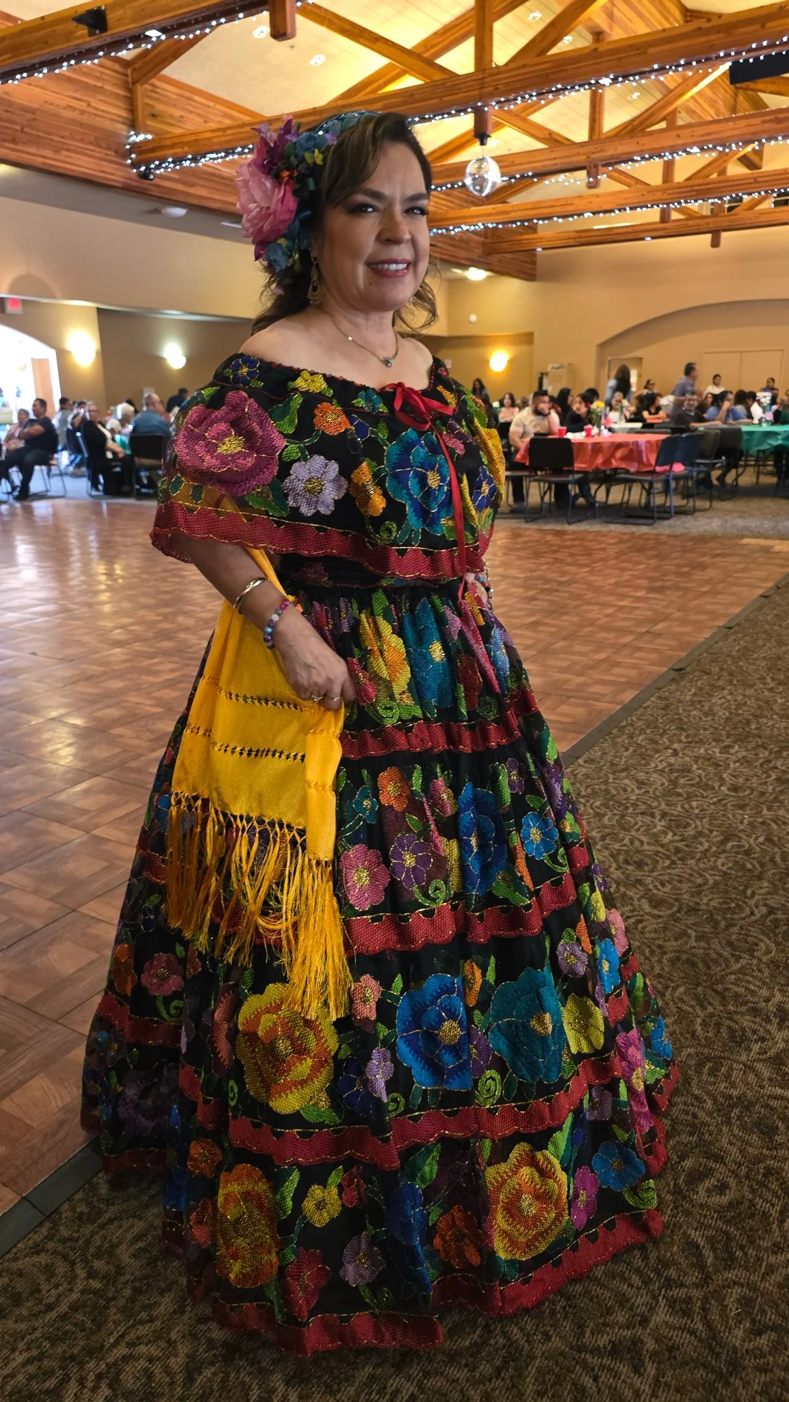 Woman wearing a traditional Mexican embroidered dress with colorful floral patterns, accessorized with jewelry and a large floral headpiece, at a festive indoor event.