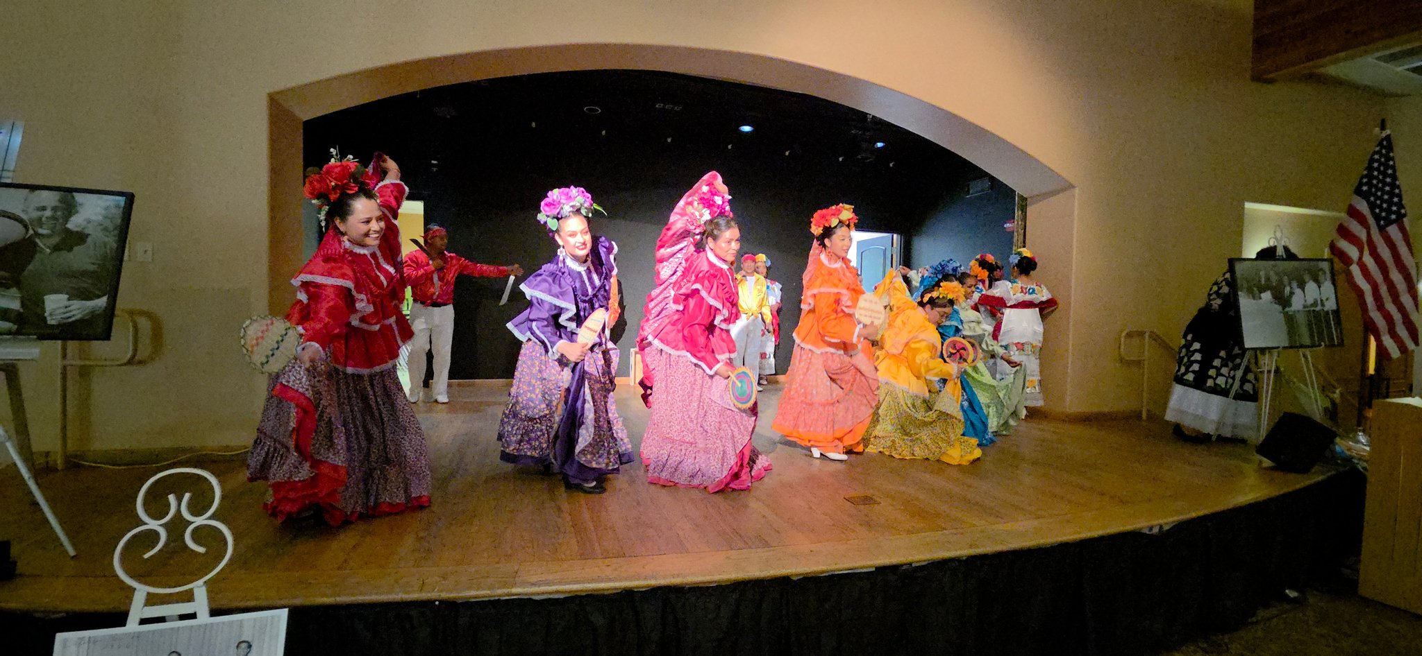 Women in colorful traditional dresses performing a dance on stage, wearing flowers in their hair.