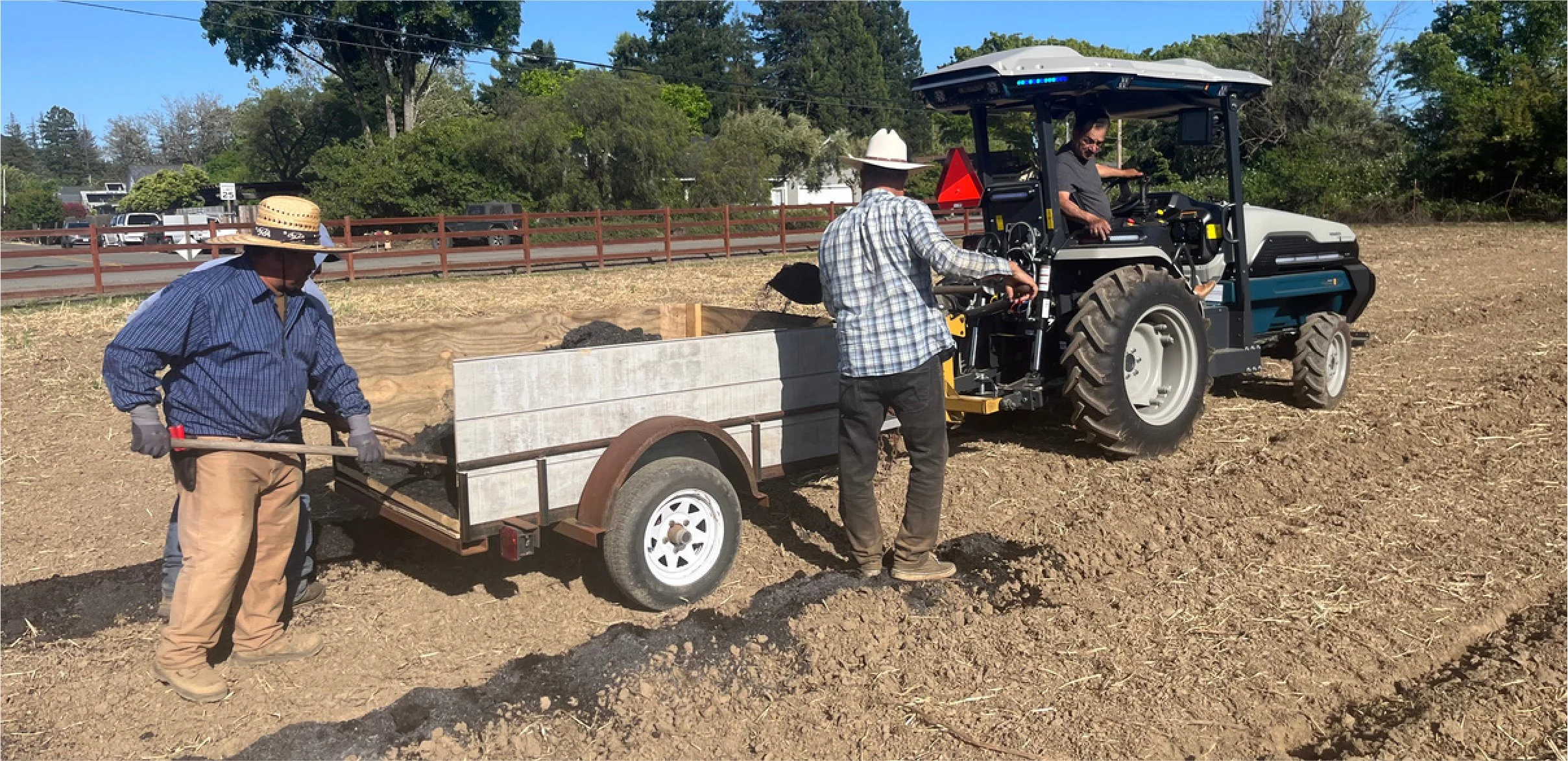 Three men working outdoors with a tractor and trailer in a dirt area surrounded by trees, with a road and car traffic in the background.