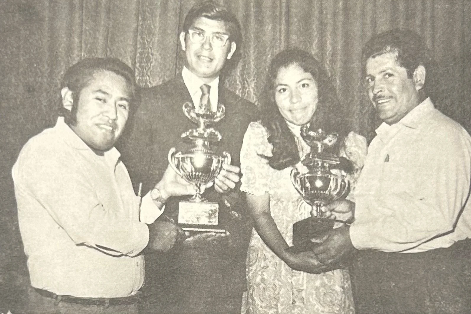 Black and white photo of four people standing together, holding trophies amidst a curtain backdrop, with two women in the center and two men on either side.